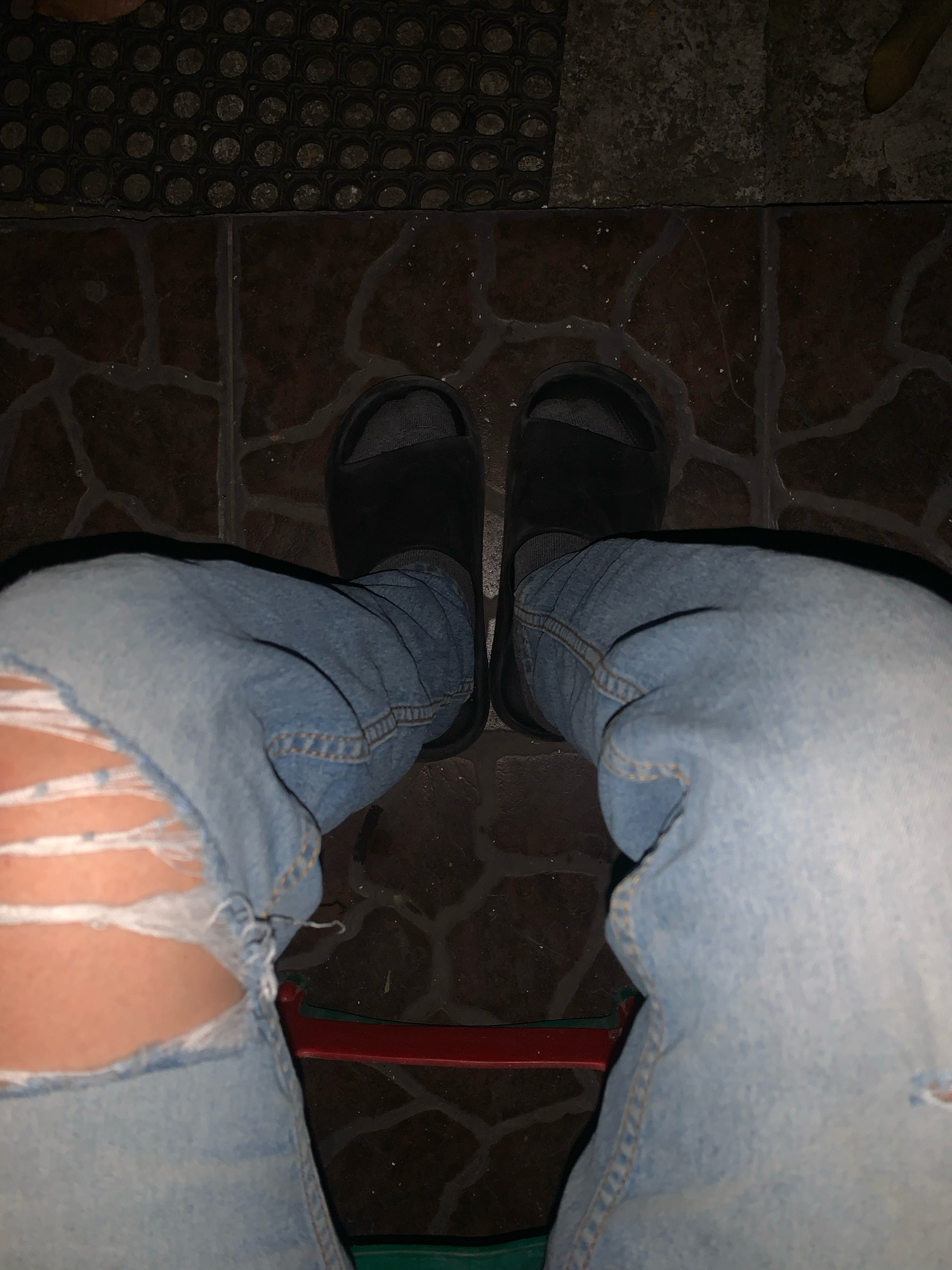The image shows a close-up view of a person's legs from the knees down, suggesting they are sitting. The person is wearing ripped light-wash denim jeans and black slide sandals over grey socks. The background consists of a textured floor with dark brown tiles and a black mat with a honeycomb pattern. A red and green object, possibly part of a chair, is visible below the jeans. The lighting is dim, with shadows cast on the floor, indicating an indoor or nighttime setting. The image does not contain any specific landmarks or elements that would allow for its localization in Sidoarjo, Indonesia.
