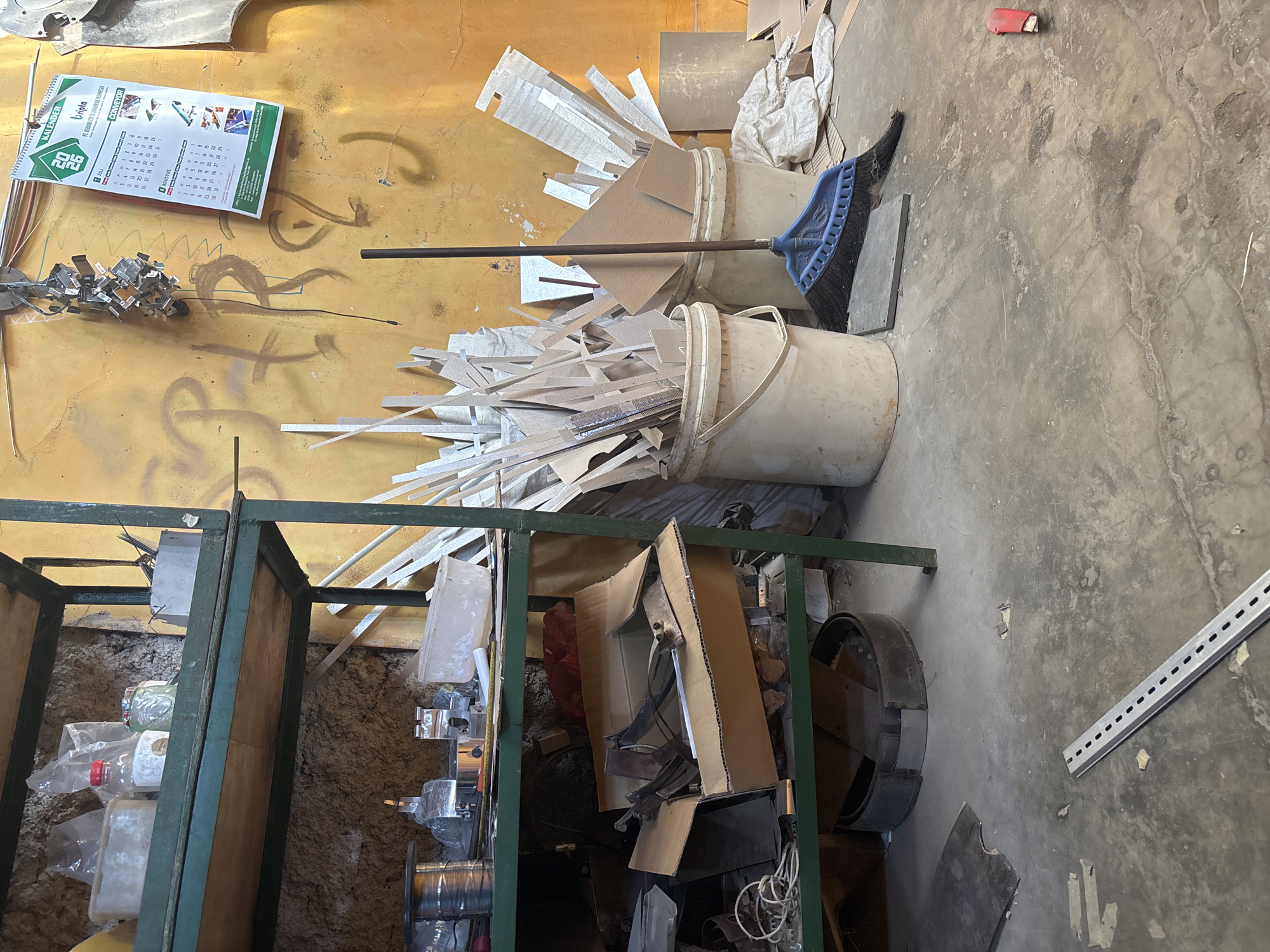 The image depicts an indoor workshop space, likely in Telagamurni, Indonesia, characterized by a cluttered and industrial environment. The scene is dominated by a green metal shelving unit filled with various items. The top shelf contains plastic containers, a glass jar, and a box of metal fasteners. The lower shelf is more disorganized, with spools of wire, metal components, and an open cardboard box containing tools and metal parts.
In the background, a yellow-painted wall is covered in graffiti. A calendar displaying July and August is hanging on the wall, alongside a cluster of small metal objects suspended from a wire. Various wooden and cardboard scraps are piled around two large white buckets. A blue broom with black bristles is positioned in front of the buckets, resting on the concrete floor.
The floor itself is a rough, grey concrete surface, showing signs of wear and tear, with scattered debris and a metal ruler lying diagonally across it. A red utility knife is visible on the floor in the lower right corner. The overall impression is of a functional, albeit untidy, workspace where manual labor and fabrication likely take place.