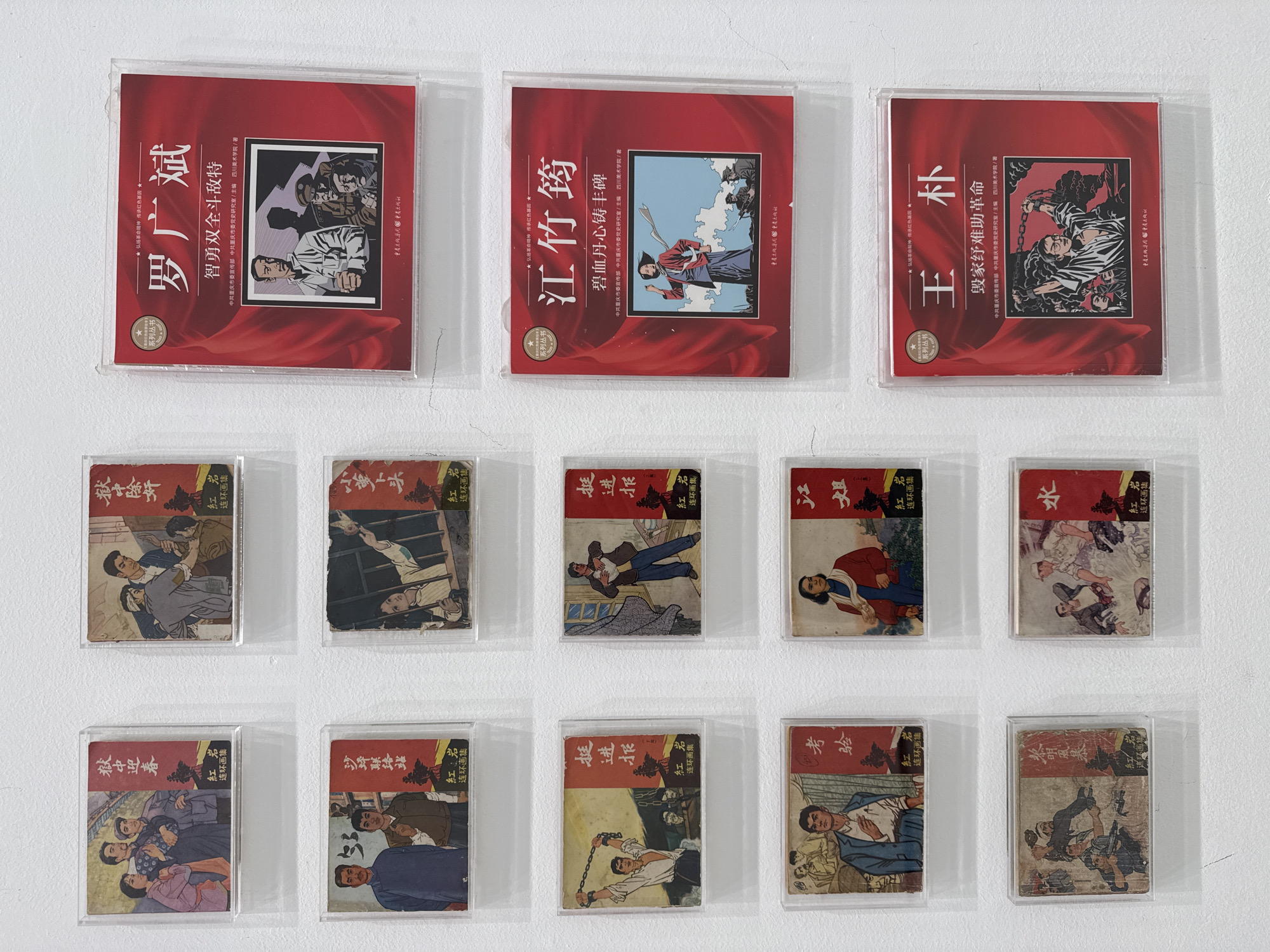 The image shows a collection of books or CDs arranged on a white wall. The items are grouped into three rows. The top row consists of three larger items, with red covers featuring illustrations and Chinese characters. The middle and bottom rows each contain five smaller items, which also appear to be books or booklets with illustrations and Chinese characters on their covers. The overall arrangement is symmetrical. Based on the OCR of the text, the setting is likely related to the city of Chongqing. The text mentions, "中共重庆市," which translates to "CPC Chongqing." Therefore, it is possible to infer that these books or CDs are related to CPC Chongqing, possibly displayed in the High-tech Zone of Chongqing, China. The books seem to be historical.