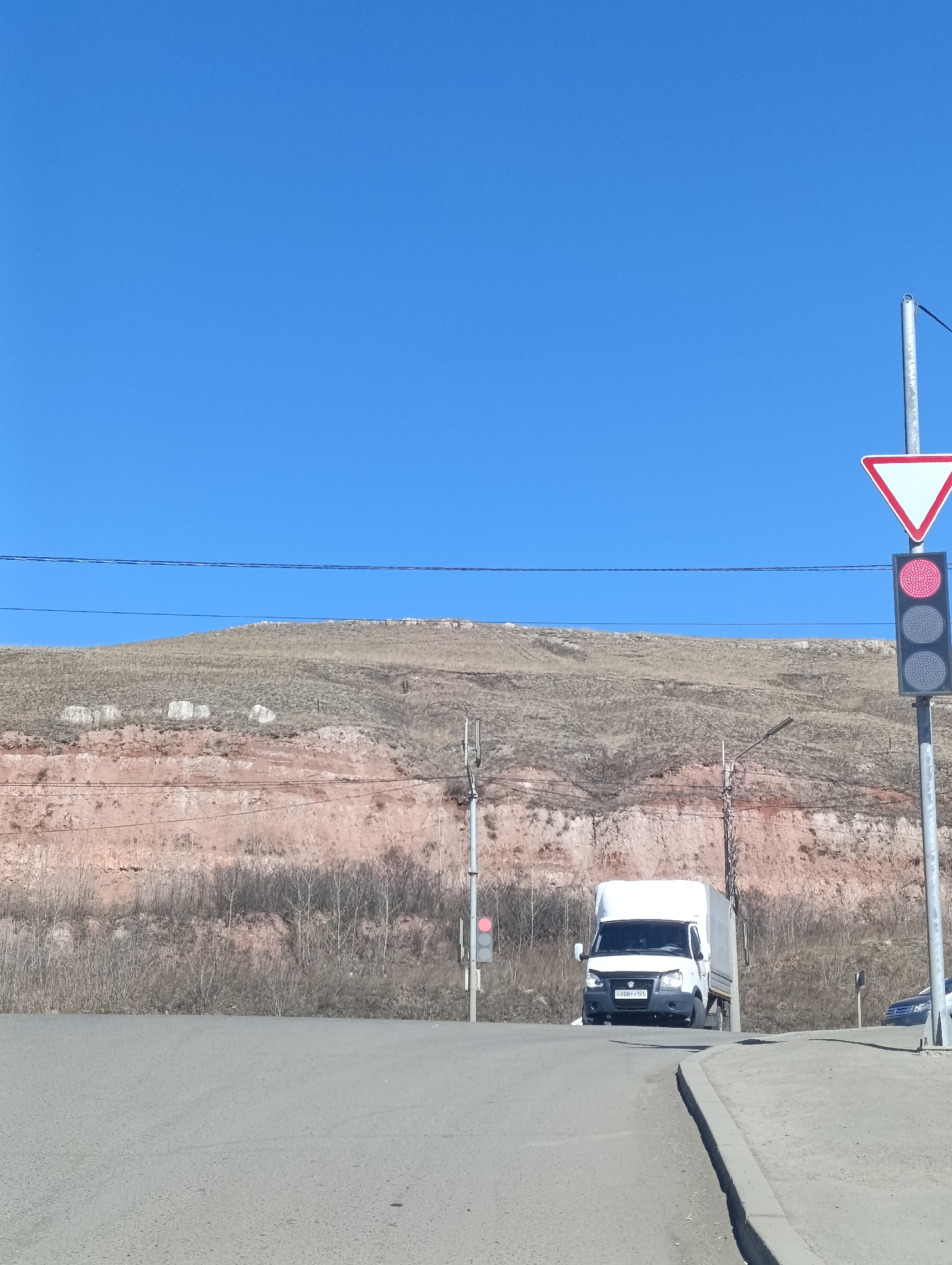 The image depicts a road in Krasnoyarsk, Russia. A white cargo van with license plate number Y208YY124 is stopped at an intersection with a traffic light showing red. A yield sign is visible next to the traffic light. A hillside with exposed reddish-brown rock strata and sparse vegetation forms the backdrop. Overhead power lines stretch across the scene. A gray car is slightly visible to the right of the van. The sky is clear and blue.