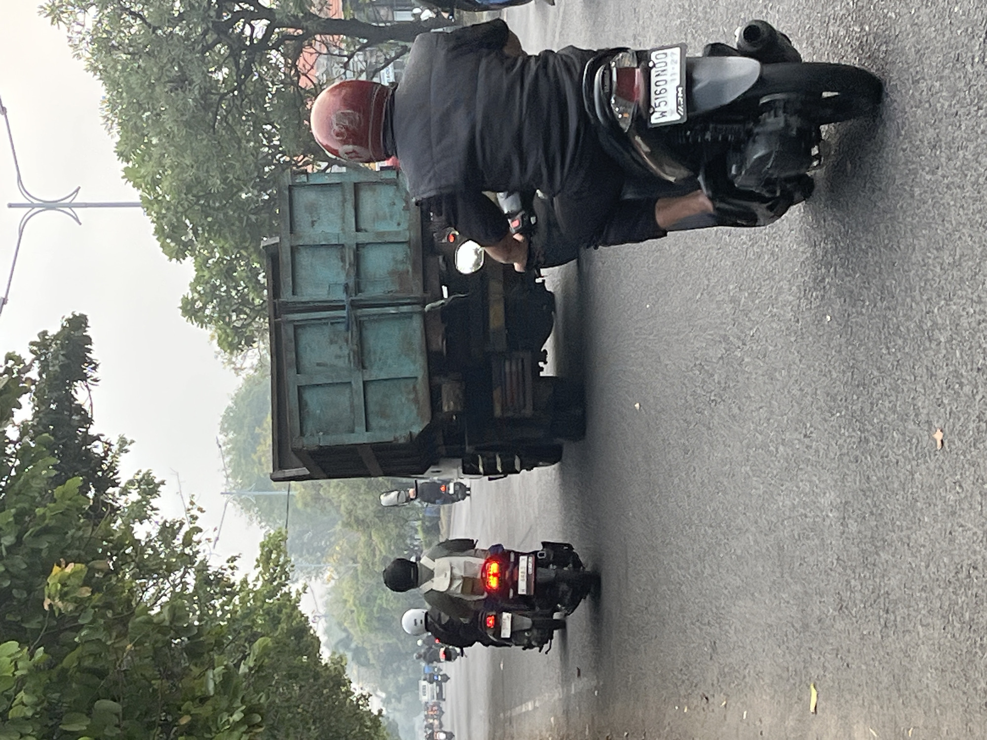 The image shows a street scene in Sumurame, Indonesia, on a hazy day. In the foreground, a person wearing a black vest and black pants is riding a black scooter, identified by the license plate "W 5160 NDO". They are wearing a red helmet. To the left, another person on a scooter is visible, wearing a white helmet and a grey jacket, with a light-colored backpack. Behind them, more scooters and a truck are on the road. Trees line the left side of the road, and a street light is visible in the background. The road surface is asphalt.