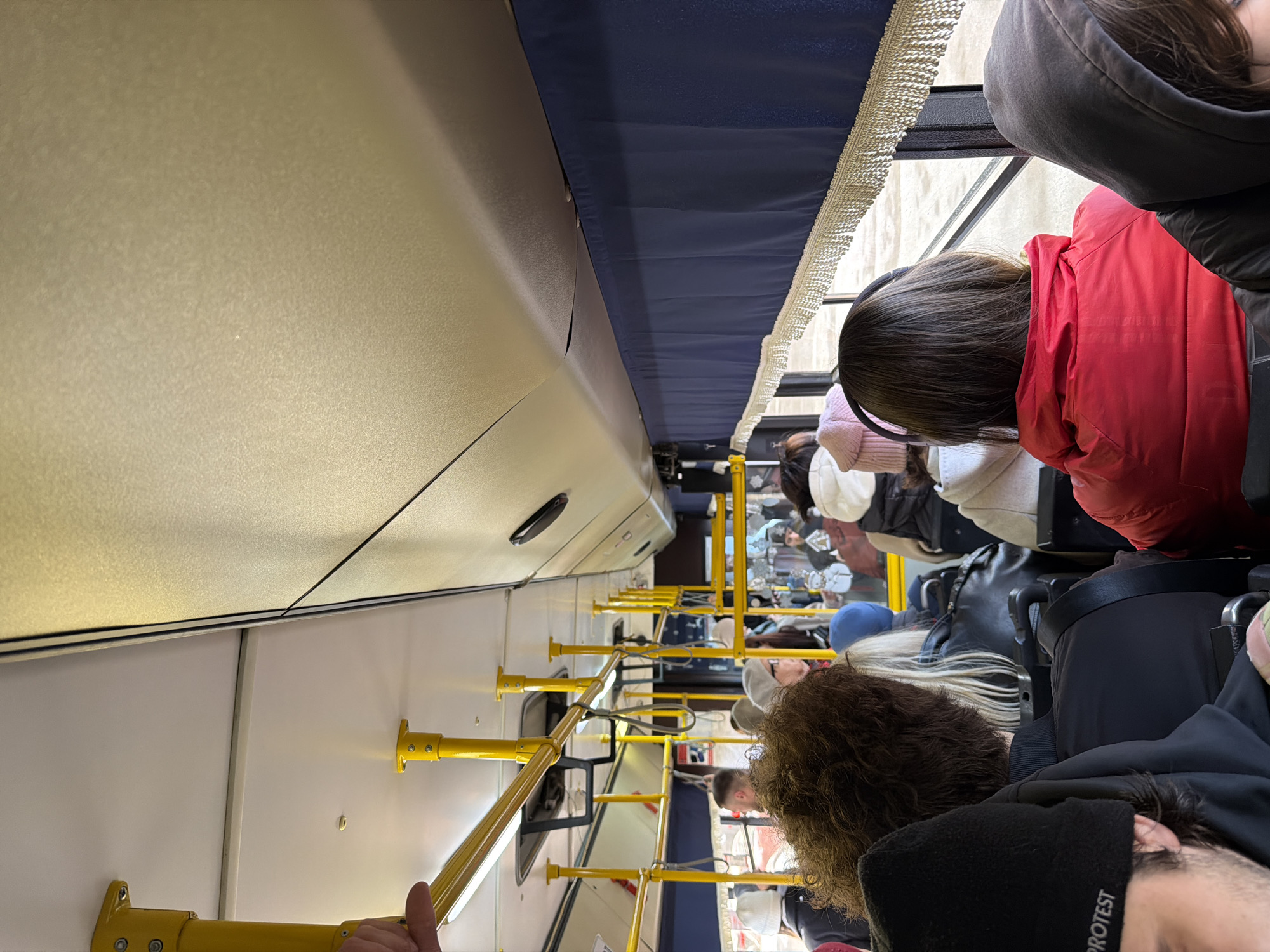 The image shows the interior of a bus, likely in Krasnoyarsk, Russia, based on the presence of Cyrillic script on signage in the background. Passengers are seated and standing, holding onto yellow handrails. Some passengers are wearing winter clothing, including hats and jackets. The interior of the bus is white with blue trim along the top. Windows are visible, revealing an urban environment outside.