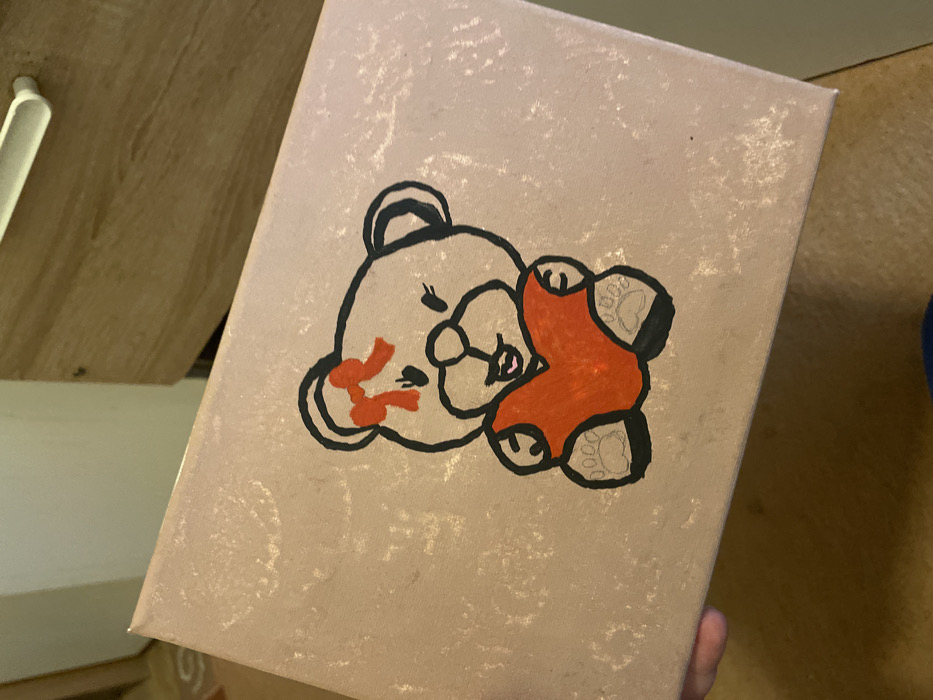 A close-up image depicts a rectangular object, likely a canvas or box lid, held by a human hand in an indoor setting. The object's surface is a speckled, light beige hue. Dominating the center is a hand-drawn illustration of a teddy bear, outlined in black, with a light beige body. The bear features a red bow on its head and holds a solid red heart in its paws. Its face includes two small black eyes, a black nose, and a smiling mouth with a pink inner detail. The paws display black pads with four lighter outlines suggesting toes. A partial human hand, showing a thumb and index finger, grips the lower-left edge of the object. The background is out of focus, revealing a light brown wooden surface with a white horizontal drawer pull in the upper right. Other blurred areas suggest indistinct interior elements. The scene is located in the city of Senden, Germany.