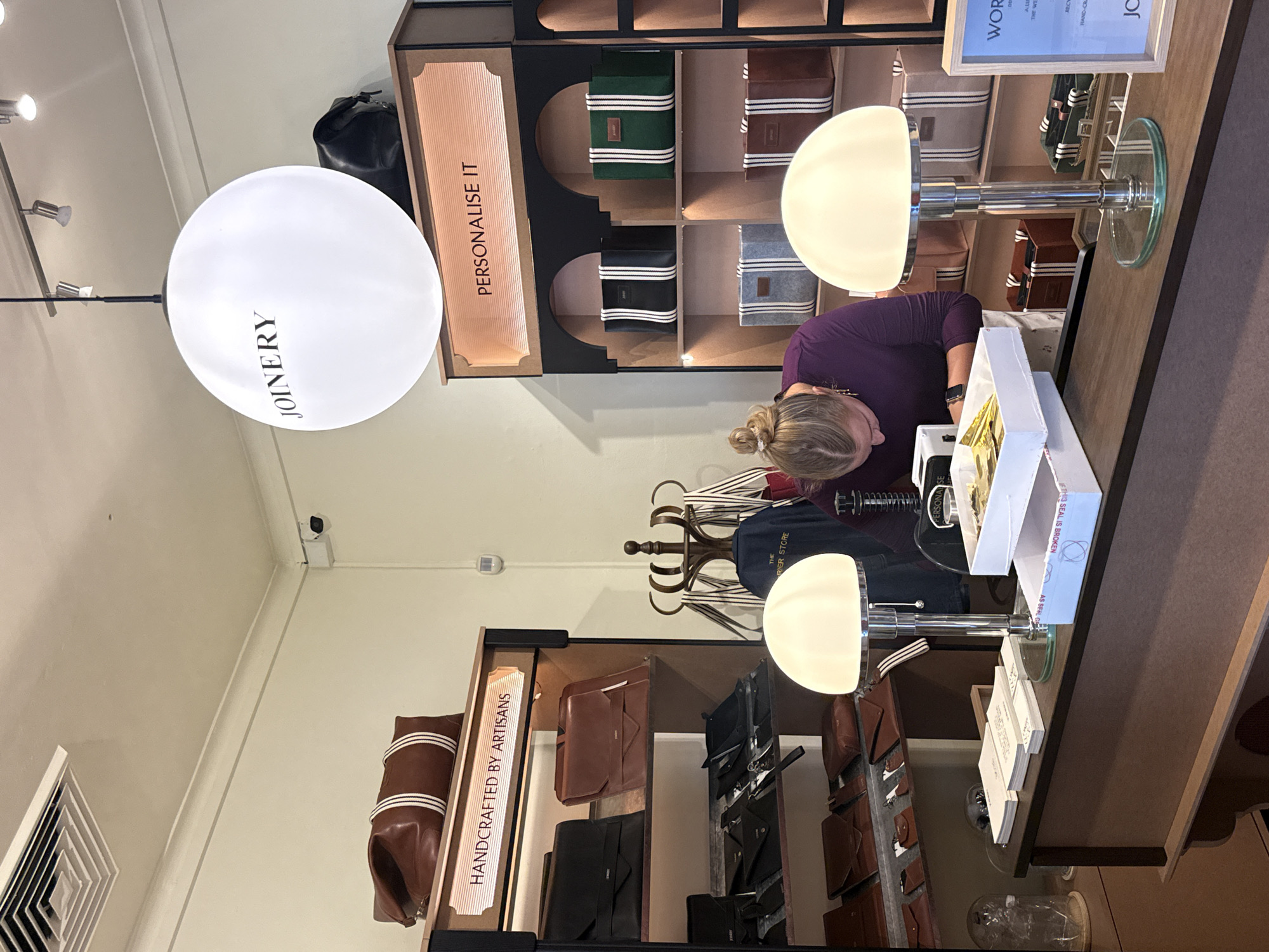 The image shows a retail store setting, specifically a store with the name "JOINERY," potentially located in Cape Town, South Africa (based on the reference to the 'RIER STORE'). A woman with blonde hair is seen inspecting items near a counter. Behind her and to the left, a rack displays various leather goods and accessories such as bags and wallets, with a sign reading "HANDCRAFTED BY ARTISANS". To her right, other shelves display similar items with a sign indicating "PERSONALISE IT". The environment features a modern and well-lit interior with white walls and spotlights on the ceiling, emphasizing the handcrafted and personal nature of the products. There are also two desk lamps on the counter.
