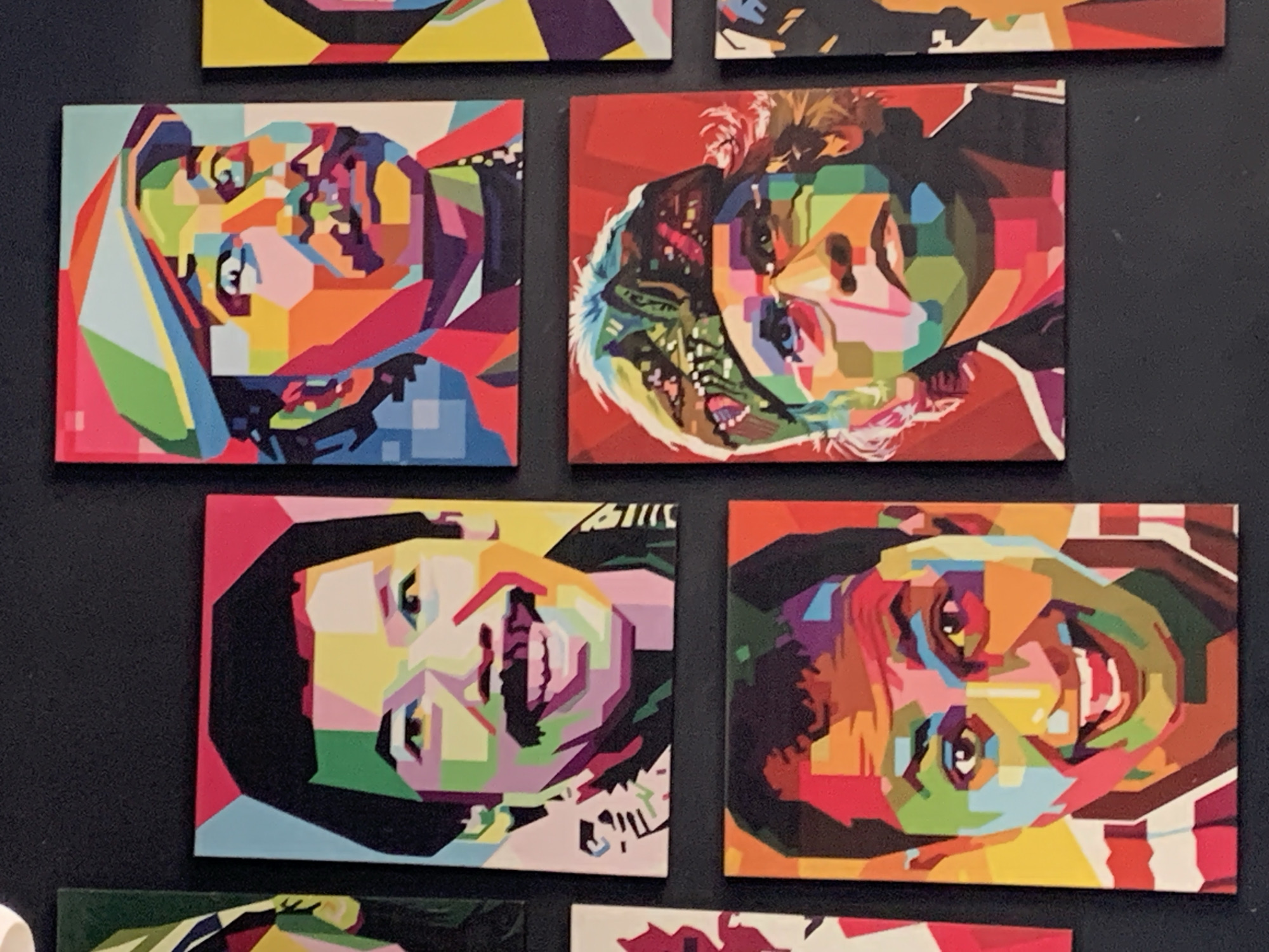 This is a medium shot, eye-level, full-color photograph of a wall adorned with several pop-art style portraits of men. The portraits are arranged in a grid-like fashion, with some pieces larger than others. The portraits depict the men in a stylized manner, using bold geometric shapes and a vibrant, contrasting color palette. The background of the photograph is a dark, textured surface, likely a wall. The lighting is even and illuminates the artwork clearly. There are no people or actions depicted in the image, only the artwork itself. The location of this artwork in Palohlada, Indonesia, is not discernible from the image itself.