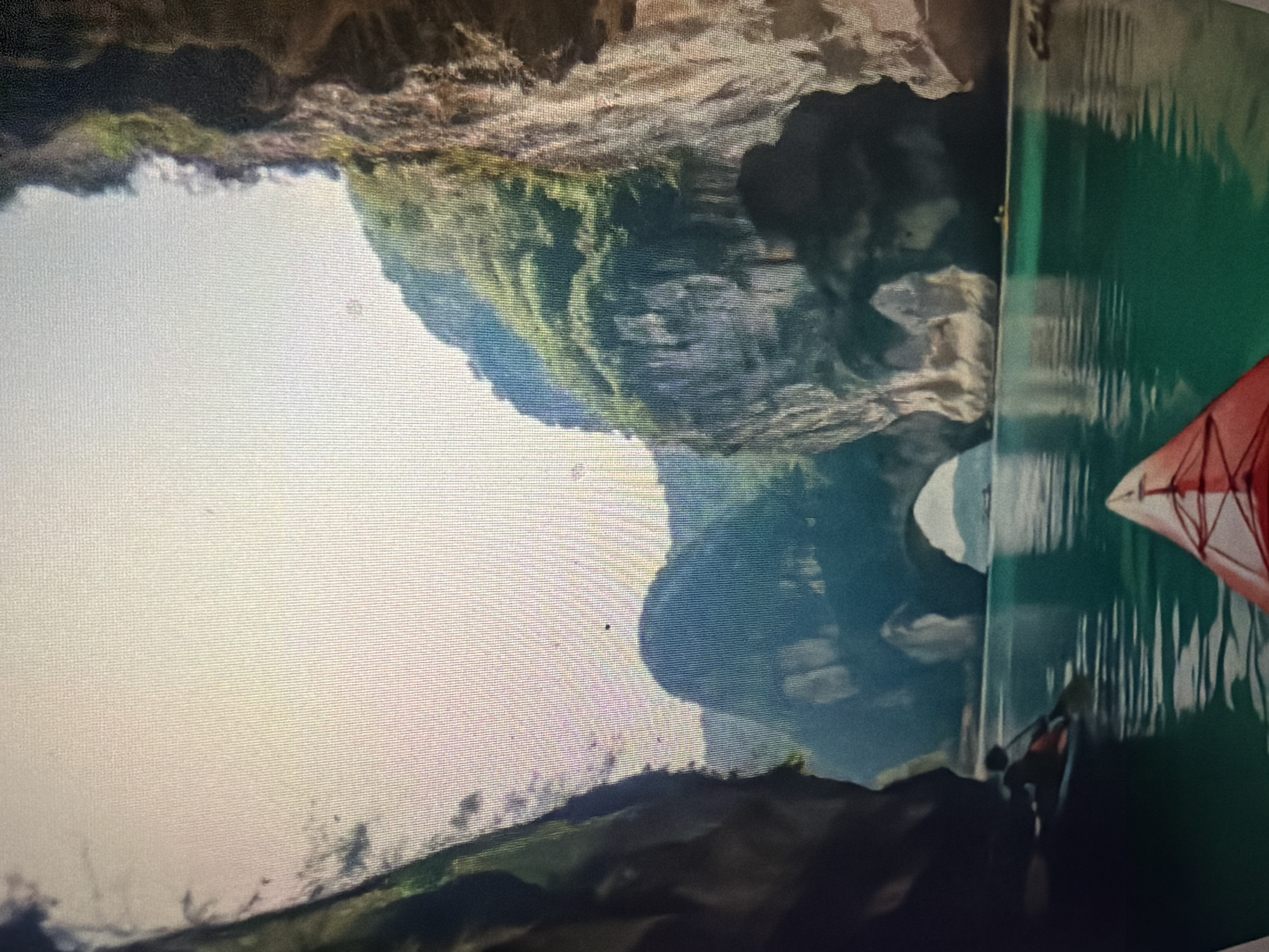 An aerial view captures a tranquil body of water, likely a bay or inlet, reflecting the soft light of the sky. Towering limestone cliffs, carpeted with lush green vegetation, flank the water, creating a dramatic natural landscape. A prominent natural archway is visible in the distance, formed by erosion through one of the cliffs, allowing passage between sections of the waterway. In the foreground, a bright red kayak is partially visible, indicating its position on the water and the perspective of the viewer. Further into the scene, a single kayaker navigates the water, their paddle dipping into the turquoise depths. The overall impression is one of serene natural beauty and adventure. The location is identified as Baan Mae Nam, Thailand.