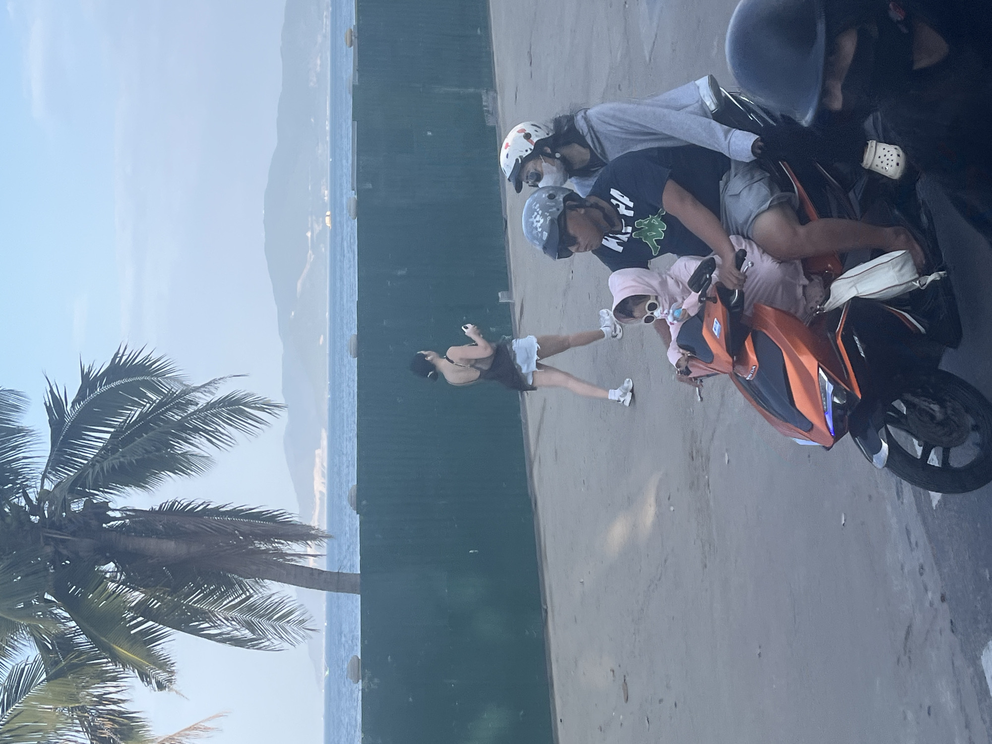 In Nha Trang, Vietnam, along a waterfront promenade, a family of four rides a motorcycle. The lead rider, a man, wears a black t-shirt and grey shorts, his head protected by a helmet. Beside him, a woman also wears a helmet and a grey hooded sweatshirt, her face partially obscured by a medical mask. In front of them, a young child, dressed in a pink hooded outfit and sunglasses, sits between them, holding a pacifier. The motorcycle is an orange and black model. In the background, a woman walks away from the camera, wearing a black tank top and denim shorts, with a small black bag. Behind her is a green construction barrier, and beyond that, the ocean with distant hills and a clear sky. A palm tree with long fronds is visible on the left side of the image.