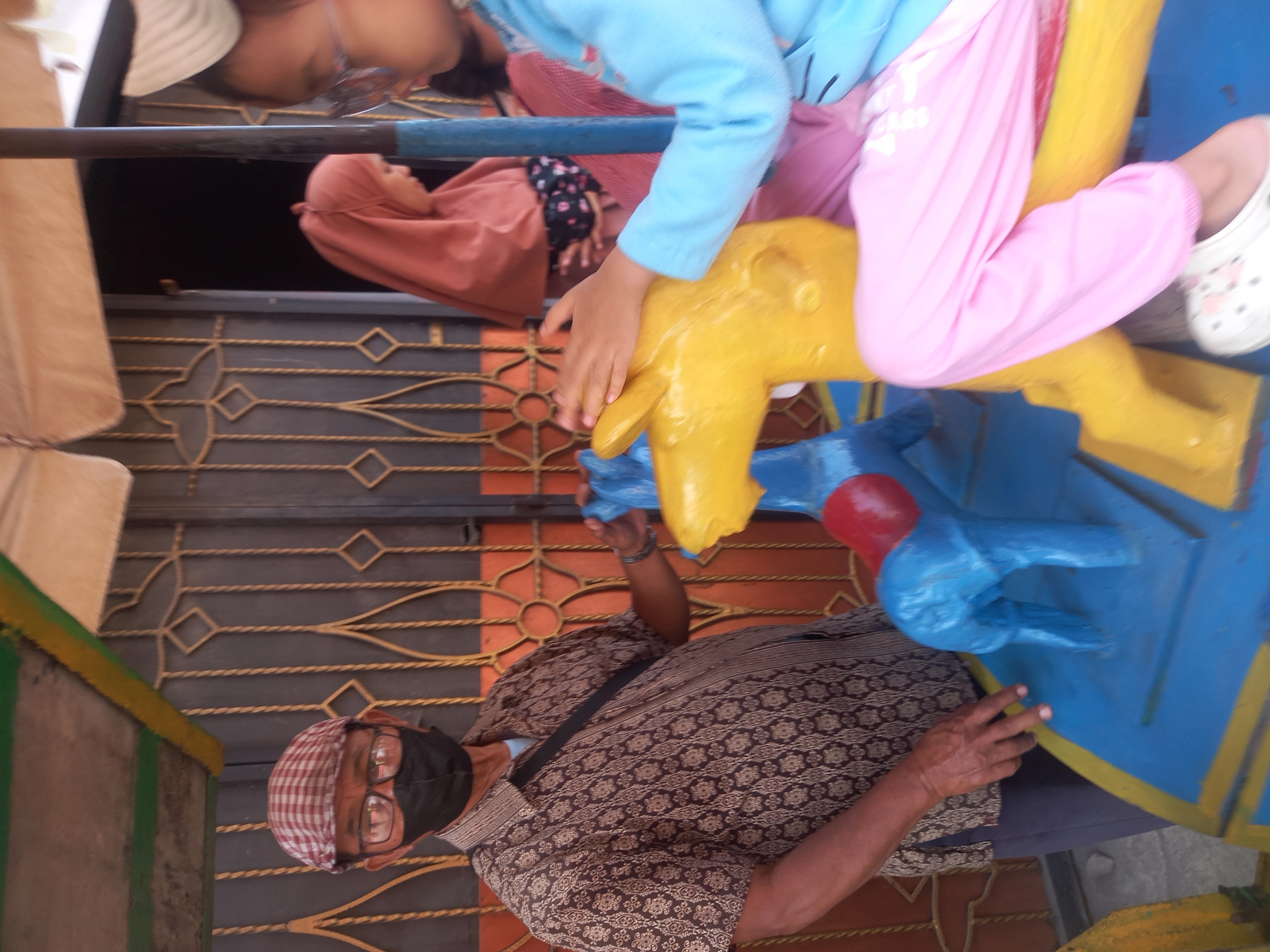An outdoor scene in the city of Linggar, Indonesia, shows a carousel with children and an adult. A man wearing glasses, a checkered cap, and a black face mask stands on the left, holding onto a blue and yellow carousel horse. He is dressed in a patterned brown shirt and dark pants. To his right, a girl in a blue shirt and pink pants is seated on a yellow carousel horse, her hand resting on its head. In the background, another person wearing a pink hijab is visible. The carousel appears to be set up in front of a building with an orange and brown patterned metal gate. The overall environment suggests a public space, possibly an amusement area or park.
