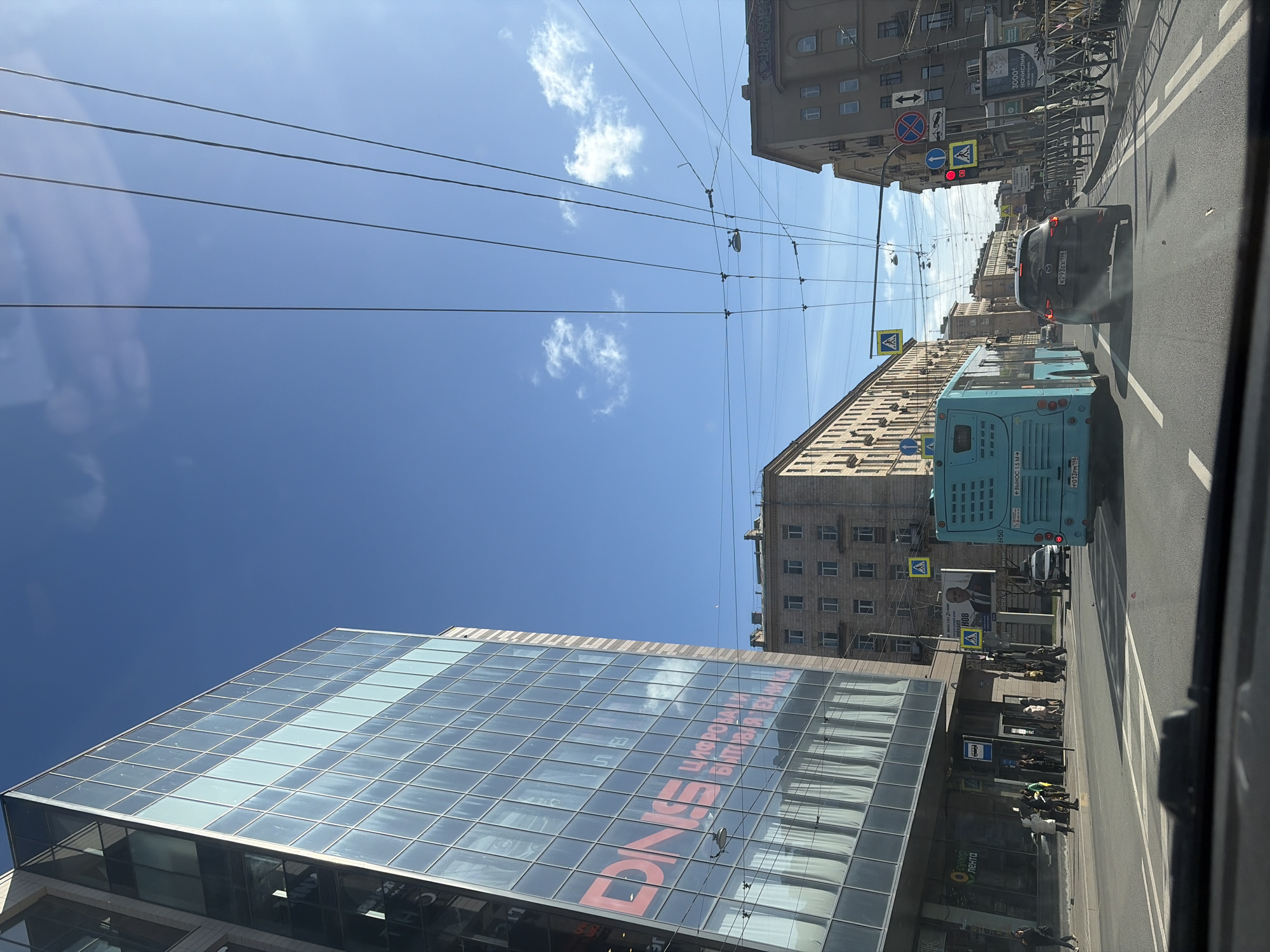 A street scene in Saint Petersburg, Russia. A modern glass-walled building with "DNS" (a Russian electronics retailer) written in red is on the left. A light blue bus travels along a road, followed by a black car. There are multiple pedestrians waiting at a bus stop. Utility lines stretch across the sky. Buildings line the street. Street signs and traffic lights are visible. The sky is clear with a few scattered clouds.