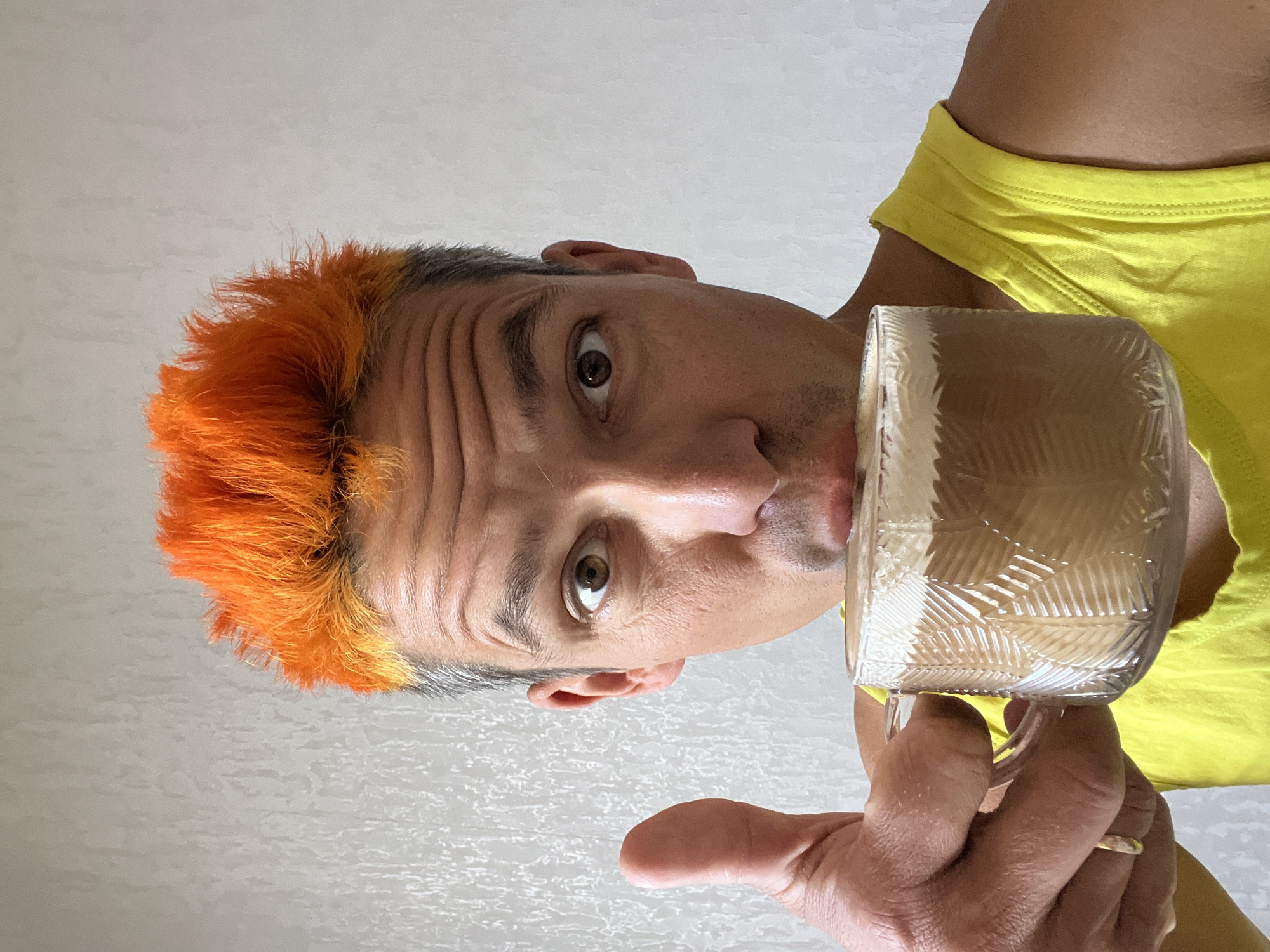 A male individual, appearing middle-aged, is depicted in a close-up, chest-up portrait within an indoor setting in the city of Chaikovsky, Russia. He features bright, spiky orange hair atop his head, transitioning to gray at the sides, and light facial stubble around his mouth. His brown eyes are open wide, and his forehead exhibits visible furrows. He wears a bright yellow sleeveless top. With his left hand, adorned with a metallic ring on the ring finger, he holds a clear, textured glass mug to his lips, from which he appears to be drinking a layered coffee beverage topped with white foam. Simultaneously, his right hand, positioned to the viewer's left of his face, forms a prominent thumbs-up gesture. He is looking directly into the camera. The background is a light-toned wall with a subtle, irregular textured pattern.