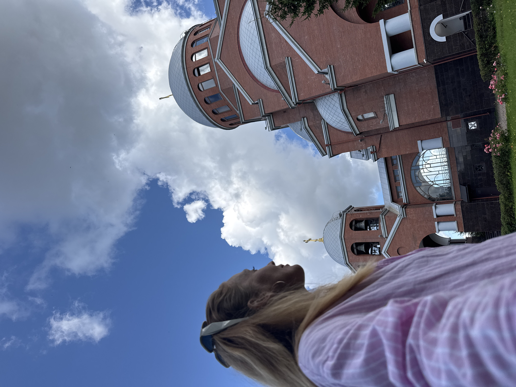 In the foreground, a woman with blonde hair, pulled back with a light blue scrunchie, is captured in profile from the chest up, facing left. She is wearing a light pink and white striped long-sleeved shirt. In the background, a large, red brick Orthodox church is visible. The church features multiple domes, some of which are silver-colored, and a prominent golden cross atop the larger dome. A bell tower with three bells is also visible on the left side of the church. The sky is blue with scattered white clouds. The church appears to be located in a green area with some rose bushes in the foreground. The architectural style of the church suggests it could be the Church of the Icon of the Mother of God of All the Afflicted, located at 103 Ligovsky Prospekt in Saint Petersburg, Russia, given its distinctive red brick construction and dome configuration, though this cannot be definitively confirmed from the image alone.