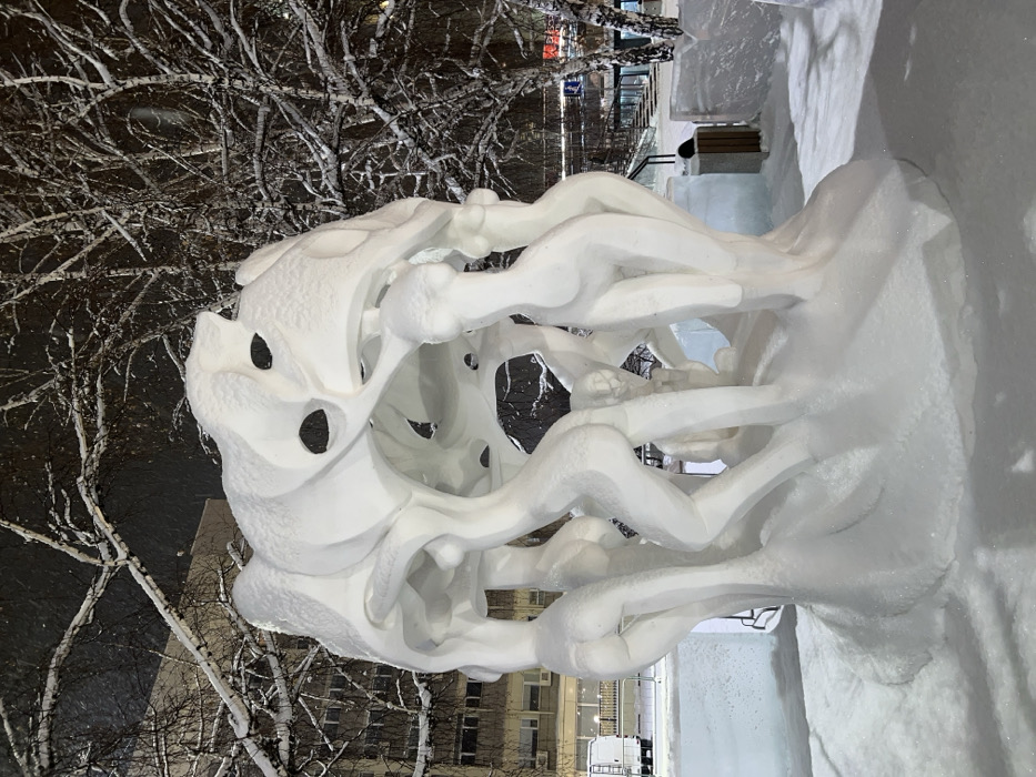 A large snow sculpture dominates a snow-covered public area in Krasnoyarsk, Russia, during a nighttime snowfall. The central sculpture features multiple human-like figures, primarily depicting female nudes in dynamic, supporting postures, holding aloft a large, abstract, organic-shaped canopy. The figures exhibit defined anatomical contours and are crafted from white snow, showing natural accumulation on their upper surfaces due to ongoing light snowfall, visible as flakes against the dark sky. The base of the sculpture is integrated into a field of undisturbed fresh snow. In the left background, a multi-story building with illuminated windows stands near a parked white cargo van. To the right, bare deciduous trees, heavily covered in snow and frost, frame the scene. Smaller blocks of ice or snow sculptures and a wooden waste bin are present on the perimeter of the main artwork. Urban infrastructure, potentially stairs or an underpass entrance, with faint artificial lighting, is visible further back on the right. The entire scene is artificially lit, creating soft shadows and highlighting the texture of the snow. The specific location is a public square or park within Krasnoyarsk, commonly used for winter art installations.