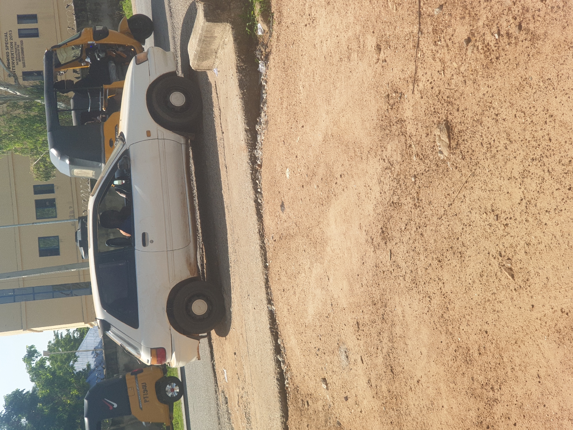 The image shows a white hatchback car and a yellow auto-rickshaw driving on a road in Jalingo, Nigeria. The car is in the foreground, and the auto-rickshaw is behind it. The road appears to be paved, with some gravel and debris on the side. There is a building in the background, and some trees. The weather is sunny. The auto-rickshaw has a registration plate that reads "PF1940". The driver of the auto-rickshaw is wearing a black shirt and a dark-colored head covering. The driver of the white car is visible inside the car, wearing a dark shirt. The car is dusty and has a black spoiler on the rear. The scene suggests a typical day on the road in Jalingo, with local transportation and buildings visible.