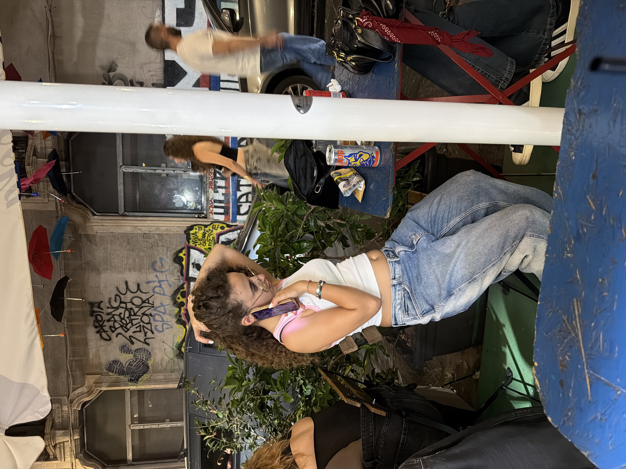 The image captures an outdoor evening scene on a street in Catania, Italy. A young woman with long, curly brown hair, wearing a white cropped tank top with pink trim and light blue jeans, is seated at a blue rectangular table, holding a purple-cased smartphone to her left ear with her left hand while her right hand is raised to her hair. She wears glasses and multiple bracelets. To her left, another woman is partially visible from the back, seated at an adjacent table.
On the blue table, a red Bull Dog energy drink can, a glass of amber liquid, and another red beverage can are visible, alongside a black shoulder bag. A prominent white pole stands centrally on the right, partially obscuring the background. In the mid-ground, a blurred woman in a black crop top and light jeans walks left-to-right, while a blurred man in a white t-shirt and blue jeans walks right-to-left behind the white pole, near a silver car.
The background features a multi-story building wall heavily covered in black graffiti, including "CAROTA BANG ROSA LATTOC ZONA SPA PIG" and "VIVA L'ATANAZ", along with other tags and abstract designs. A window with a metal grate is visible on this wall. Numerous small, inverted umbrellas in red, orange, black, blue, and pink are suspended overhead as decoration. Potted green plants are arranged around the seating area. The ground under the tables is a mix of dark pavement and a green surface. In the bottom right, the legs and feet of a person wearing dark jeans and white Adidas sneakers are visible, with a red bandana tied around one leg.