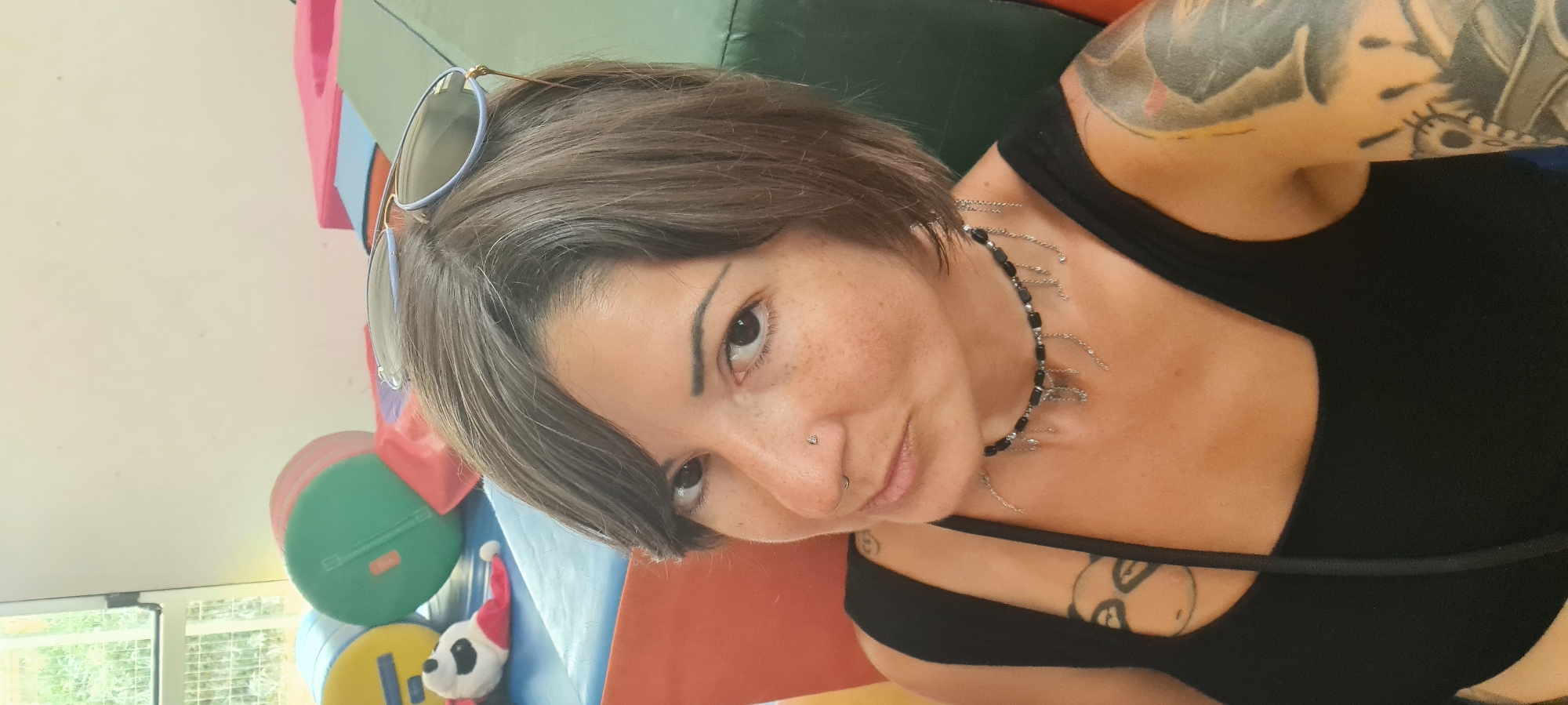 A woman with short grey hair, freckles, and a nose piercing is pictured in a selfie. She wears sunglasses atop her head and a black tank top. A silver and black necklace adorns her neck. A tattoo is visible on her left upper arm. In the background, there are colorful soft play objects, and a stuffed animal wearing a Santa hat. The environment appears to be indoors, likely in the city of Lachish Regional Council, Israel.