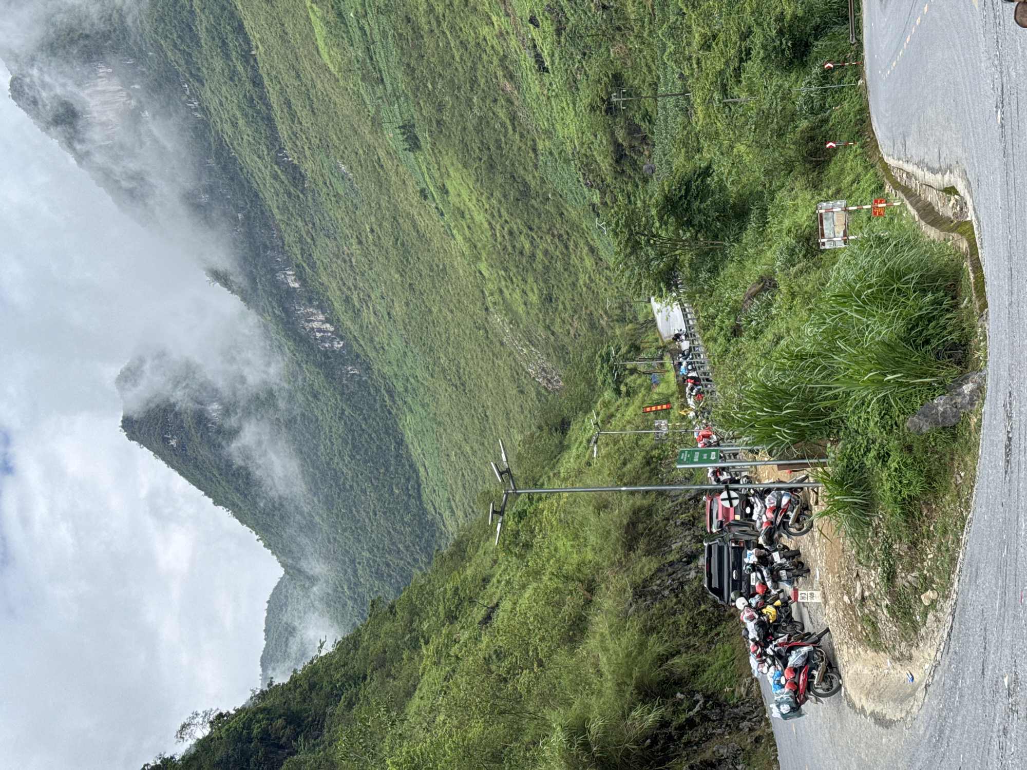 The image shows a roadside scene in the Yên Minh Commune, Vietnam, with steep, green, mountainous terrain in the background partially obscured by low-lying clouds. A narrow asphalt road curves to the right of the frame. Parked along the roadside is a dense cluster of motorcycles and at least one black pickup truck. Utility poles and signage are visible along the road. The surrounding landscape is covered in lush vegetation.