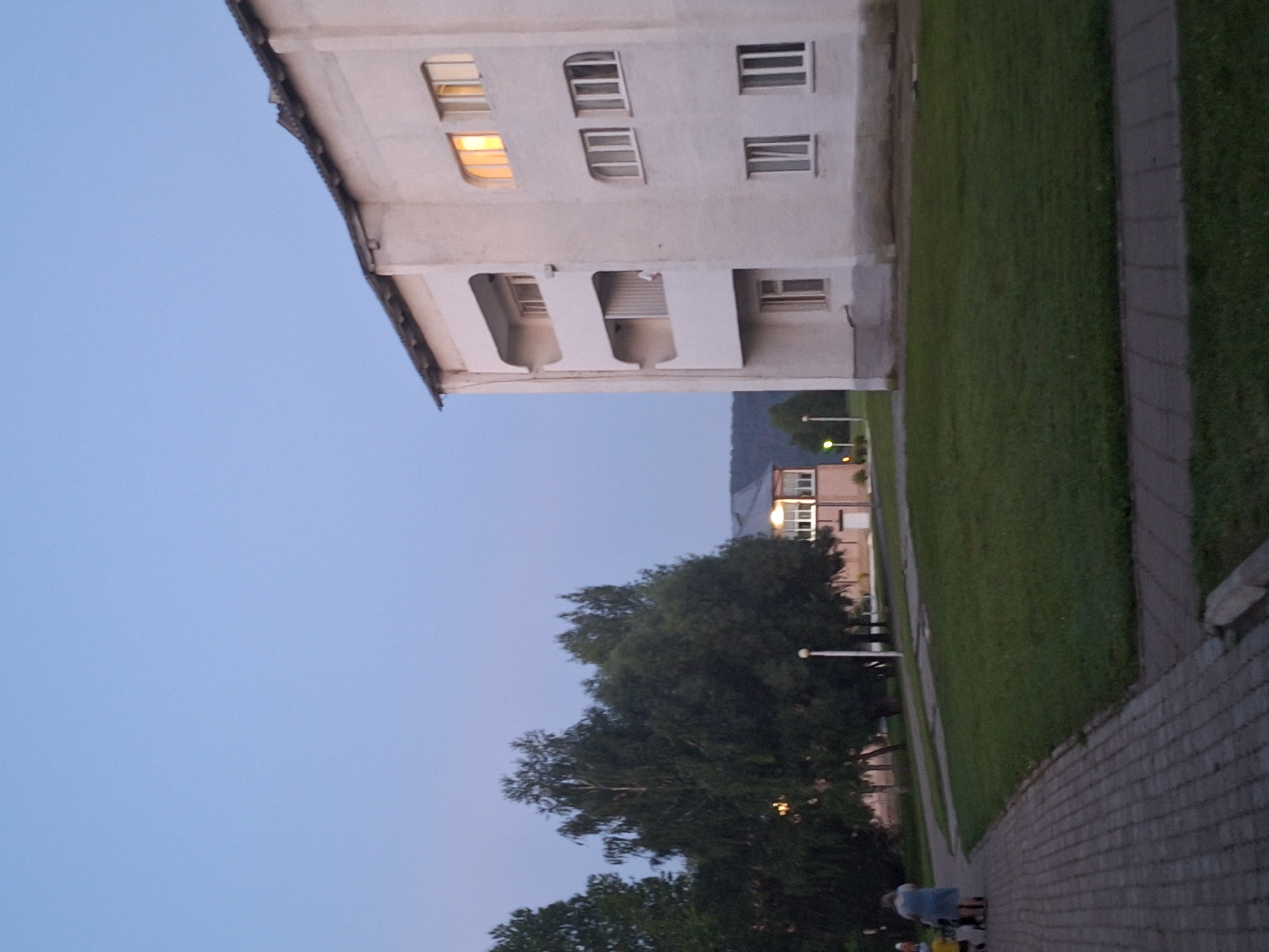 The image depicts an outdoor scene in Атамановка, Russia, at dusk. In the foreground, a paved walkway leads away from the viewer to the left. A woman in a blue dress is pushing a stroller with a child, and another child is walking beside them. To the right of the walkway is a well-maintained lawn.
Dominating the right side of the frame is a three-story building with a white facade. The building has balconies on the upper two floors and several windows. One window on the top floor emits a warm yellow light, indicating an interior light source. The roofline of this building is visible at the top.
In the background, to the left of the main building, is another, smaller building with a light-colored facade and a darker roof. This building also has windows, and one appears to be illuminated with a yellow light. A lamppost stands on the lawn in front of this building. Behind the buildings, a dense line of trees, including birch trees, creates a dark green backdrop against the pale blue evening sky. The distant landscape shows rolling hills. The overall atmosphere is peaceful and quiet, characteristic of a residential area in the early evening.