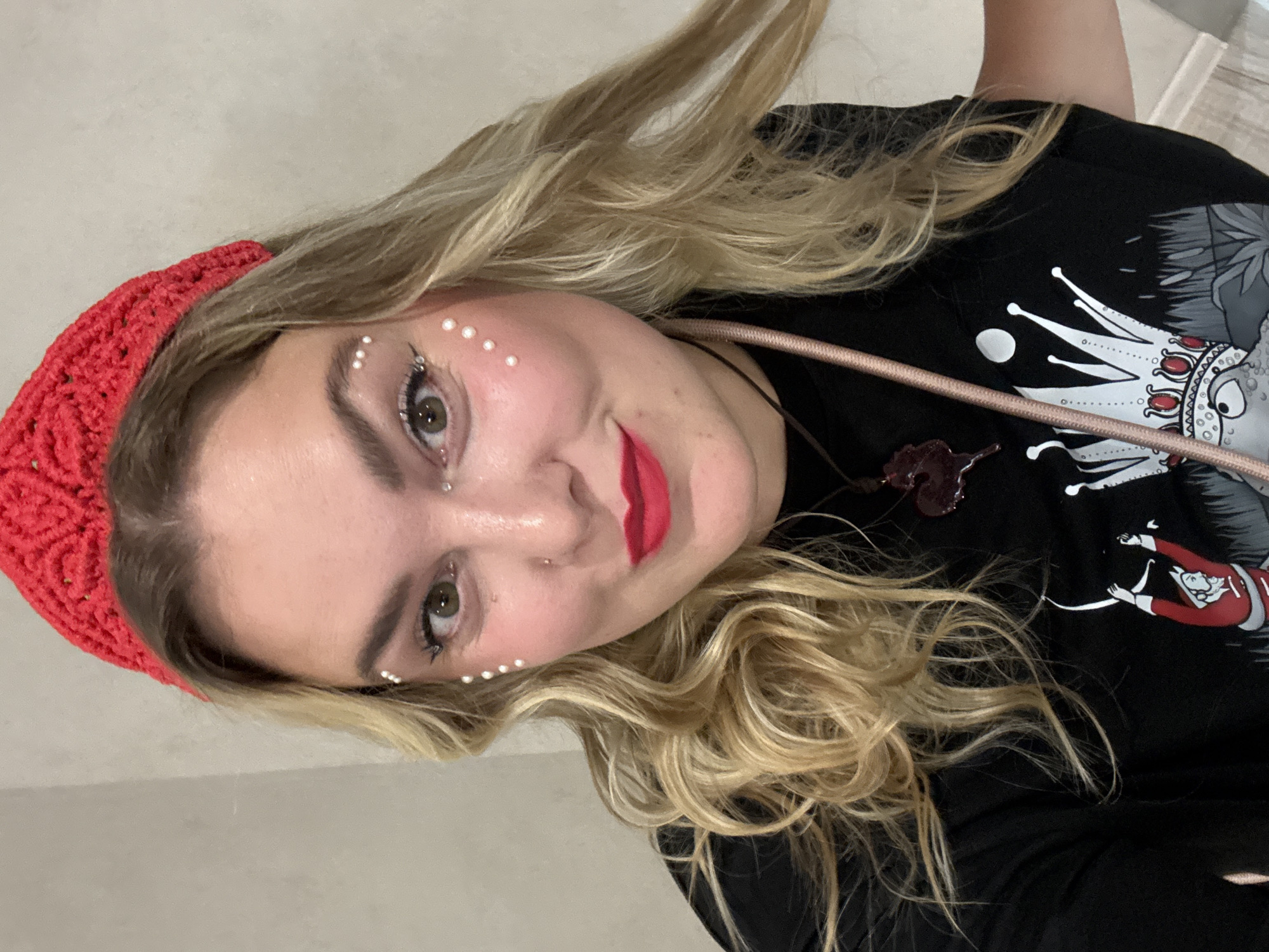 The image is a close-up, selfie-style shot of a young woman with blonde, wavy hair, wearing a red knitted headband. She has applied makeup, including red lipstick and small white pearls or beads strategically placed around her eyes and on her cheeks. She is wearing a black t-shirt with a graphic print featuring a crown and stylized figures. A rose-gold colored strap from a bag is visible across her chest. The background appears to be a plain, light-colored wall. The lighting is soft and appears to be indoors. There is no indication of the location being in the city of Kungur, Russia.