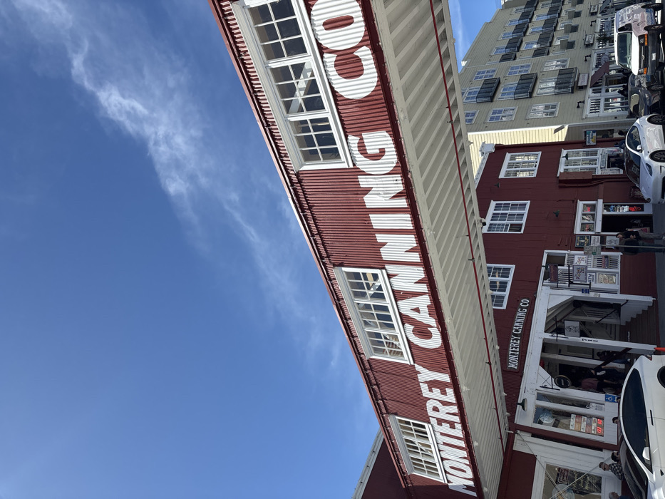 The image captures an outdoor daytime scene on Cannery Row in Monterey, California, United States. The central focus is a two-story building complex constructed primarily of dark red corrugated metal with white trim and numerous white-framed windows. An overhead pedestrian bridge, also featuring red corrugated metal, spans across the scene with "MONTEREY CANNING CO" written in large white letters. The ground floor of the main building features multiple storefronts with white wooden facades, one prominently marked "MONTEREY CANNING CO" and "711". Several individuals are visible on the sidewalk and entering the building. Parked on the street in front of the building are a white Tesla Model X (partially visible on the left), a white Tesla Model Y, a dark grey SUV, and a white van. To the right of the red building, a taller, multi-story beige building with multiple balconies and windows is visible. The sky is clear and blue with sparse, wispy white clouds.