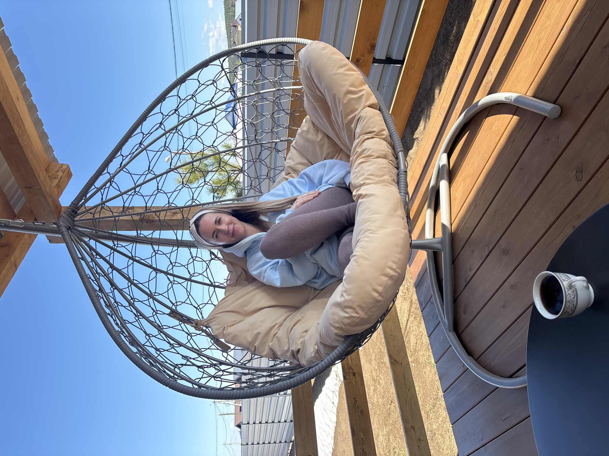 The image shows a woman sitting in a hanging egg chair outdoors. She is wearing a light blue hoodie, brown leggings, and a white headband. The chair has a beige cushion. A cup of coffee sits on a black table next to the chair. The background includes wooden decking, a corrugated metal fence, and a bright blue sky. Based on visual clues and the provided location, the scene is likely taking place on a patio or deck in Chaikovsky, Russia.
