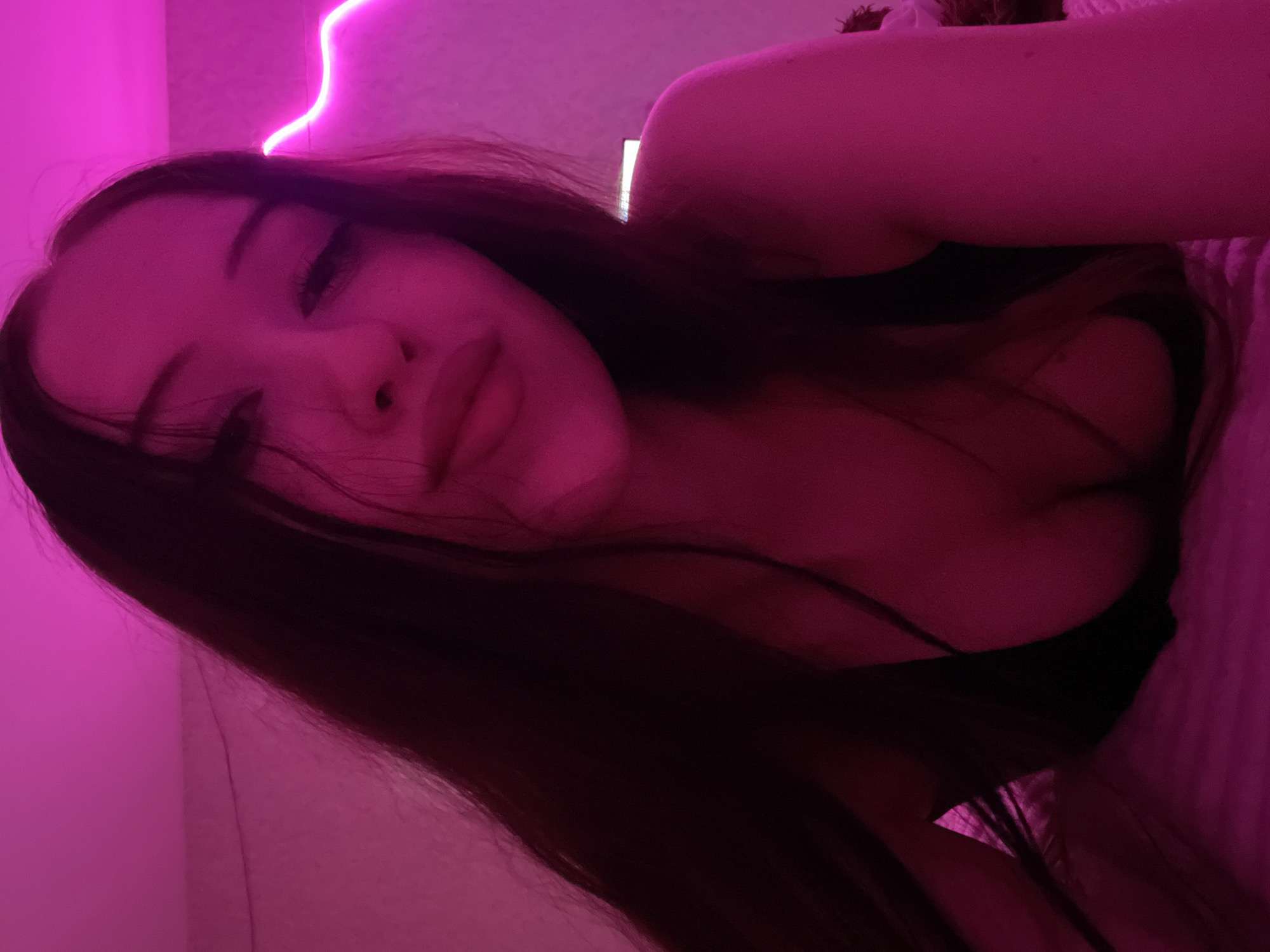 The image shows a close-up of a young woman lying down in bed. She has long, dark hair that falls around her shoulders and chest. Her head is tilted, and she is looking towards the camera with her eyes partially closed. She is wearing a dark-colored top that reveals her cleavage. The room is lit with pink neon lights, which cast a vibrant glow on her skin and surroundings. The background appears to be a wall with the neon light strip attached to it. There is no indication of her specific location within Votkinsk, Russia, beyond the general context of a domestic indoor setting. The image is an informal selfie, suggesting a personal and relaxed atmosphere.
