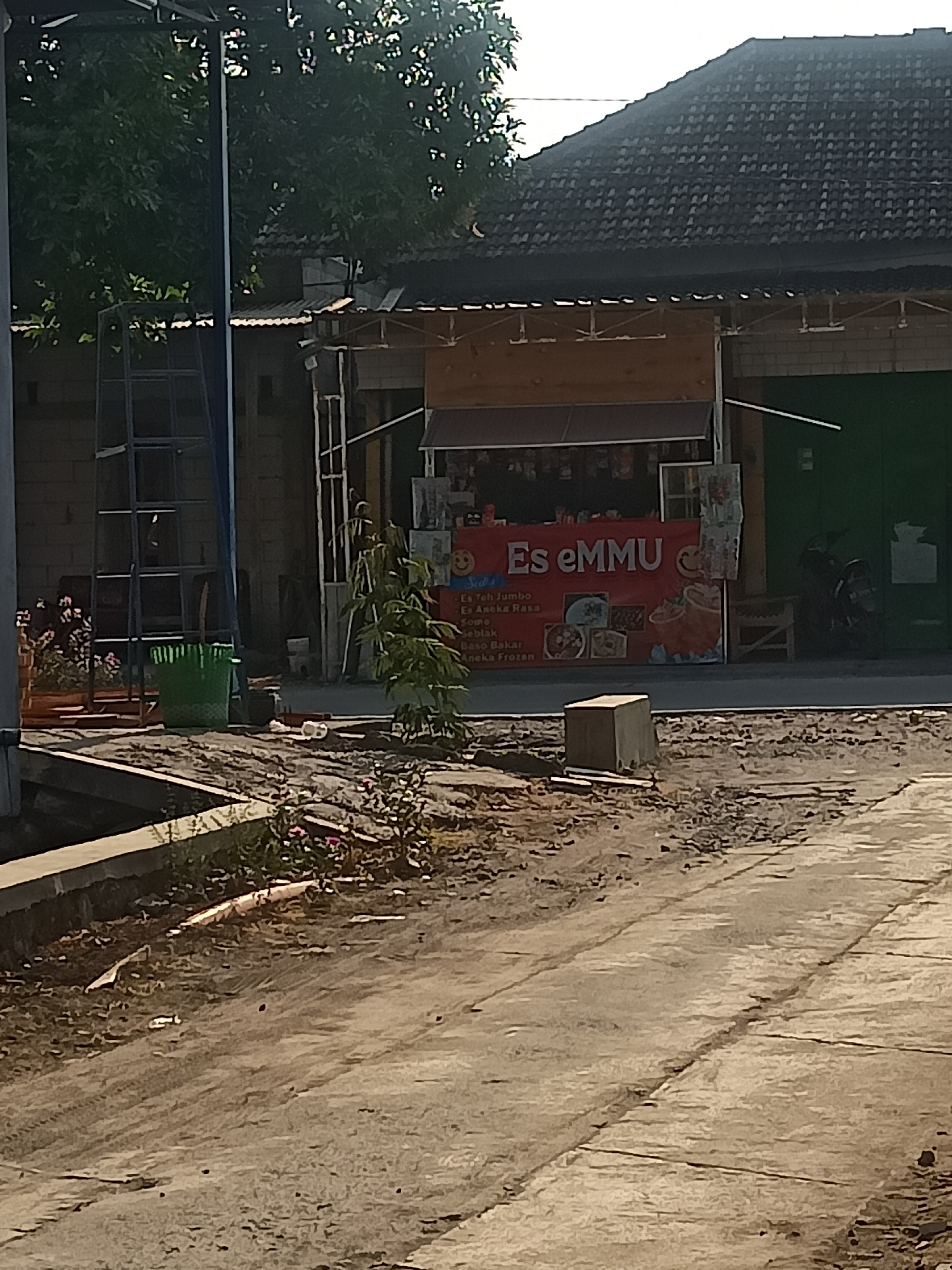 The image depicts a street scene in Karangtalun, Indonesia, featuring a small food stall or warung. A red banner with the text "Es eMMU" is prominently displayed, indicating the name of the establishment and likely its primary offering of iced beverages. Beneath this banner, a menu board lists various items including "Es Teh Jumbo," "Es Aneka Rasa," "Cumi," "Seblak," "Baso Bakar," and "Aneka Frozen," suggesting a diverse range of snacks and drinks. To the right of the stall, a motorcycle is parked. In the background, a building with a dark tiled roof is visible, along with trees and vegetation. In the foreground, a rough, unpaved area with dirt and debris is present, bordering a paved walkway or road. A green bucket and a metal structure are also visible near the stall. The lighting suggests it is daytime, likely late morning or early afternoon, given the shadows cast.