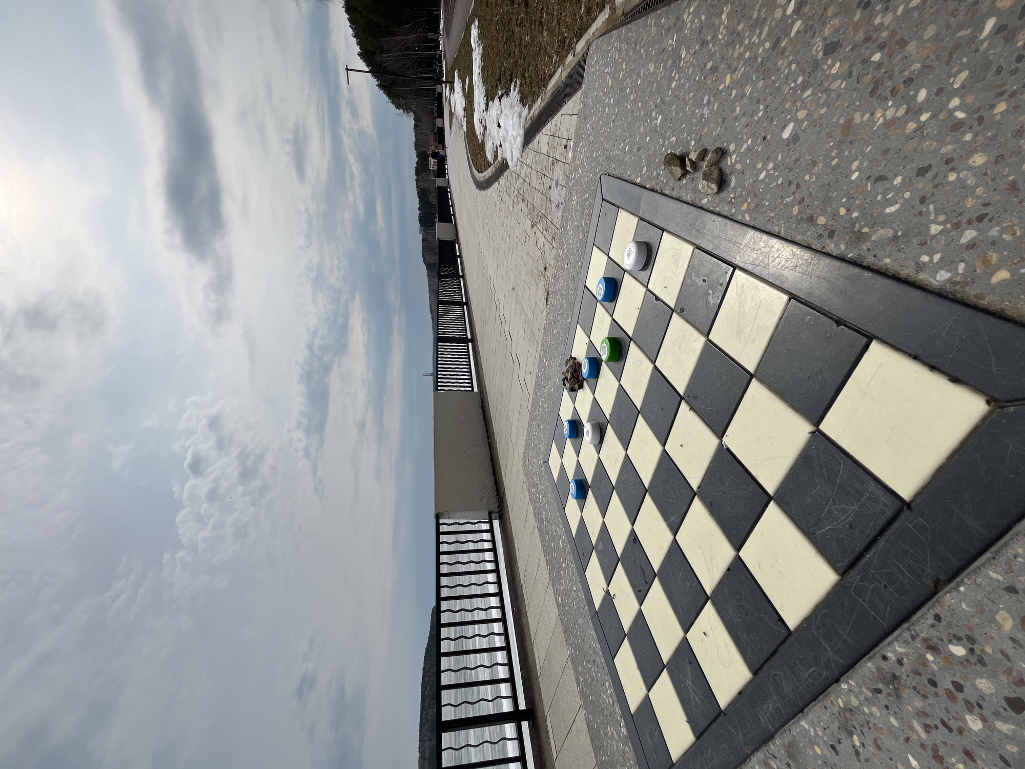 The image depicts a public space in what is likely Chaikovsky, Russia, based on visual cues such as architecture and foliage. A checkerboard is integrated into the walkway's pavement, with several checkers pieces placed on it. The pieces are various colors, including white, blue, and green. Adjacent to the checkerboard, there is a metal railing likely overlooking a lower area or body of water. The surrounding ground is paved and has a speckled concrete appearance. In the background, there is a grassy area with patches of snow, suggesting it is early spring or late winter. Beyond that, there is a line of trees. The sky is overcast, with clouds dominating the upper portion of the image.