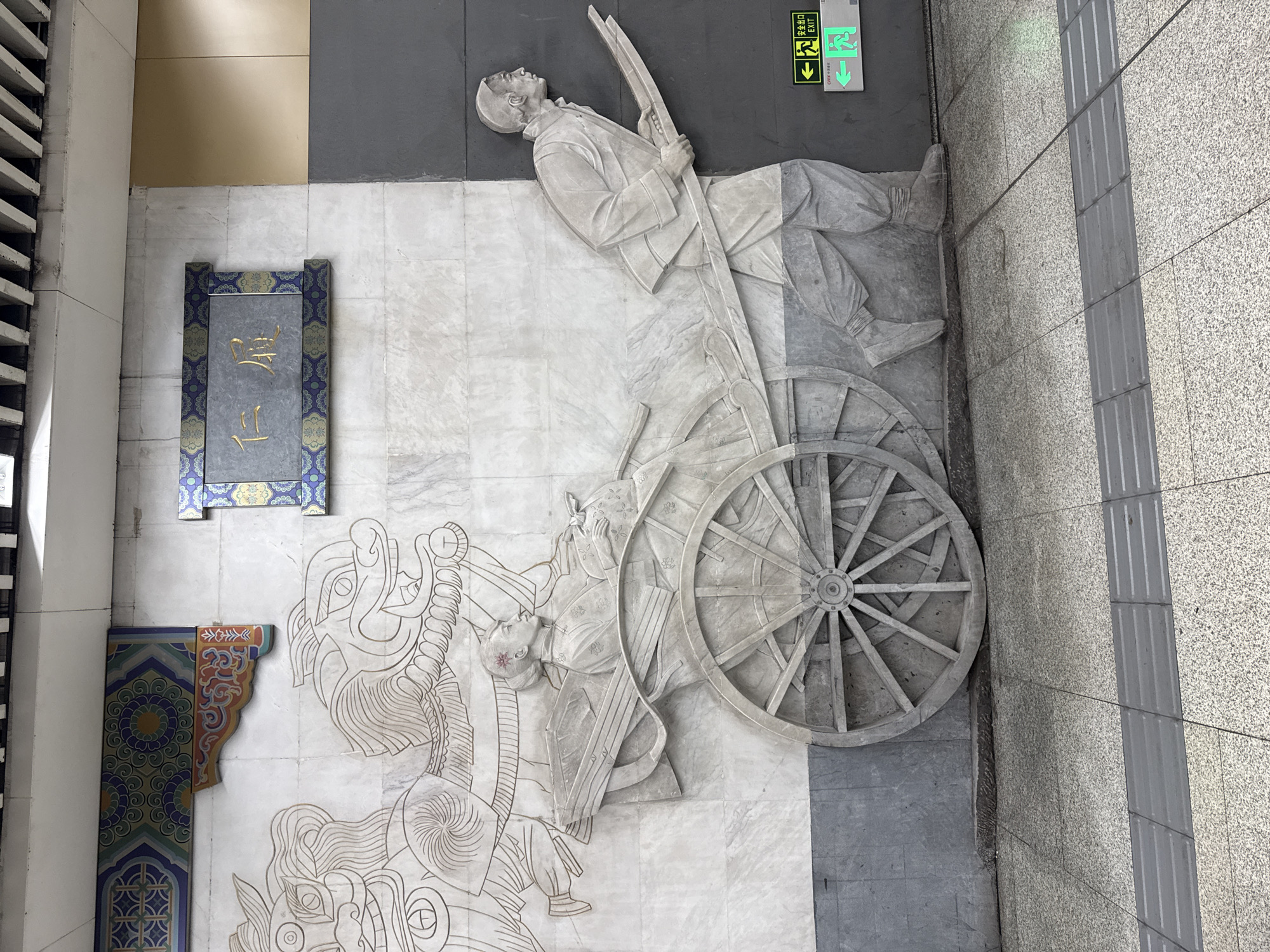 The image depicts a large bas-relief carving on a marble wall, located in Dongcheng District, China. The carving features a historical scene with two main elements. On the left, two stylized, dragon-like creatures are depicted, possibly in motion, with flowing manes. To the right of these creatures, a woman is seated in a two-wheeled cart. She is dressed in traditional attire and holds a large, round object. The cart is being pulled by a man, also dressed in traditional clothing, who is shown in profile, leaning forward as he pushes the cart's handles. The carving is rendered in a monochromatic, stone-like material against the white marble background. Above the carving, a rectangular plaque with Chinese characters and decorative borders is affixed to the wall. The environment appears to be an indoor public space, possibly a subway station or an underpass, with a grey, textured wall section to the right of the carving. Green exit signs are visible on this grey wall section. The floor in front of the carving is tiled with a speckled pattern, and a tactile paving strip runs parallel to the wall.