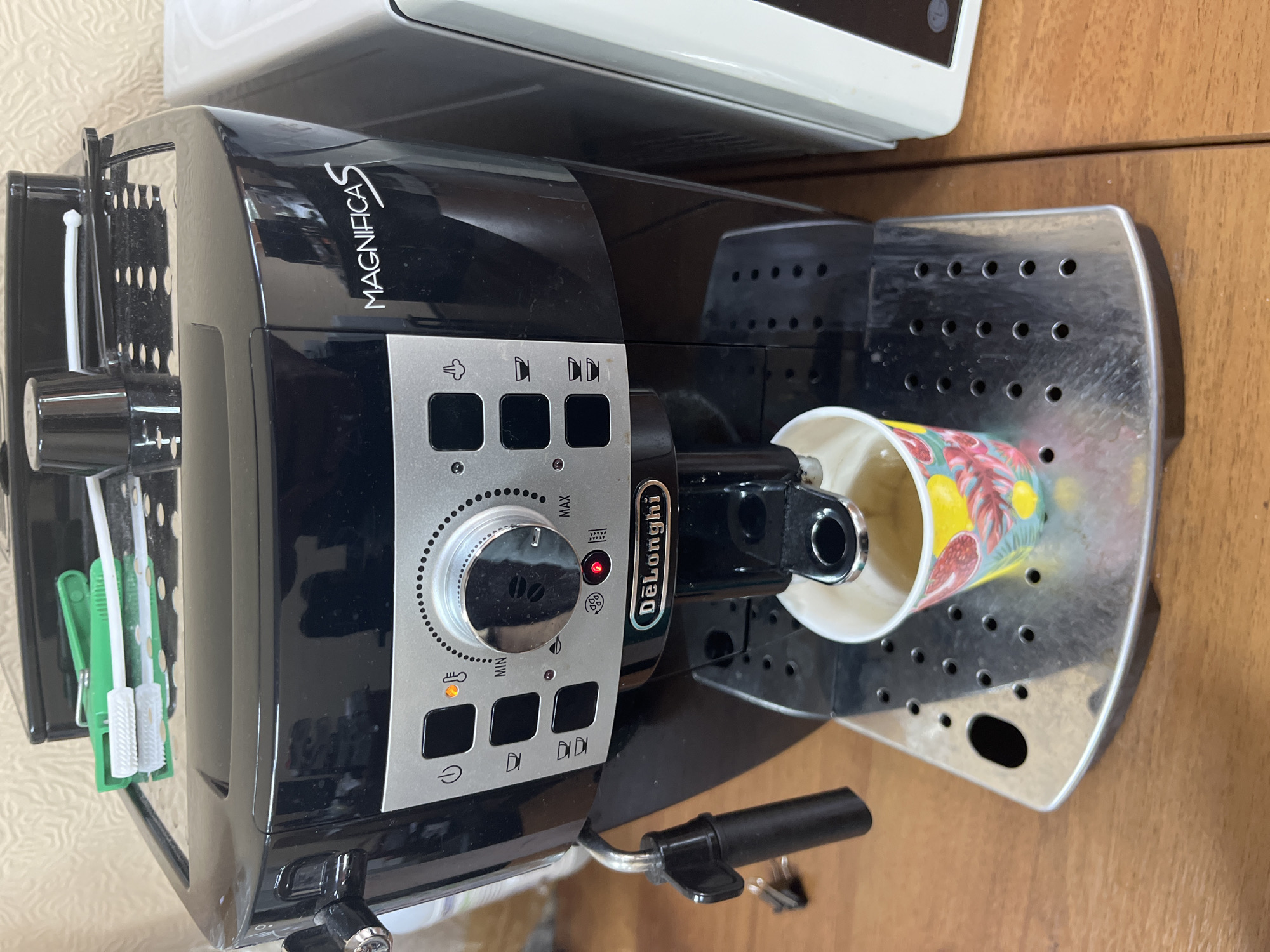 A black DeLonghi MagnificaS espresso machine is shown on a wooden table. A cup of coffee is being brewed into a colorful paper cup. The machine has a control panel with buttons and a dial. There are also two green cleaning brushes on top of the machine. The scene appears to be indoors, with a textured wall in the background. There is no indication of the city of Sochi, Russia in the image.