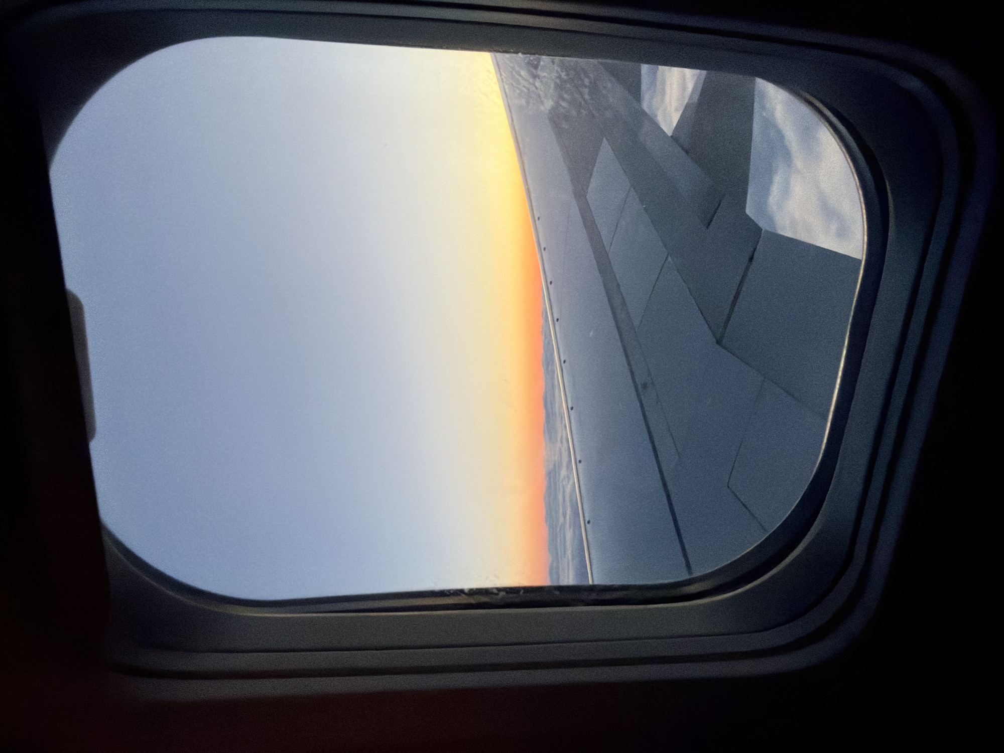 The image captures a serene view from an airplane window, framed by the dark, rounded-rectangular interior of the aircraft. The scene outside depicts a breathtaking atmospheric phenomenon, indicative of either sunrise or sunset.
The left side of the visible sky is a gentle, pale blue-grey, which gradually transitions into a vibrant gradient of yellow, orange, and pink hues towards the right, creating a distinct horizon line. This colorful band suggests the sun is low on the horizon, bathing the sky in warm tones.
Occupying the right side of the window is a portion of the airplane's wing. It appears metallic grey, with visible panels, seams, and what might be rivets or other structural details along its surface. There are also some subtle textures on or reflecting off the wing, possibly frost or very thin clouds. The wing extends from the right edge of the window, angling slightly upwards and backwards.
The dark frame of the window is prominent, especially in the top-left and bottom-left corners, where the interior structure of the plane is visible, emphasizing the view through the pane. The overall impression is one of peaceful flight during dawn or dusk. No specific activity beyond observation from the plane is shown, and no people are visible. There is no discernible text within the image. The flight is occurring in the general location of Lokorsko, Bulgaria.
