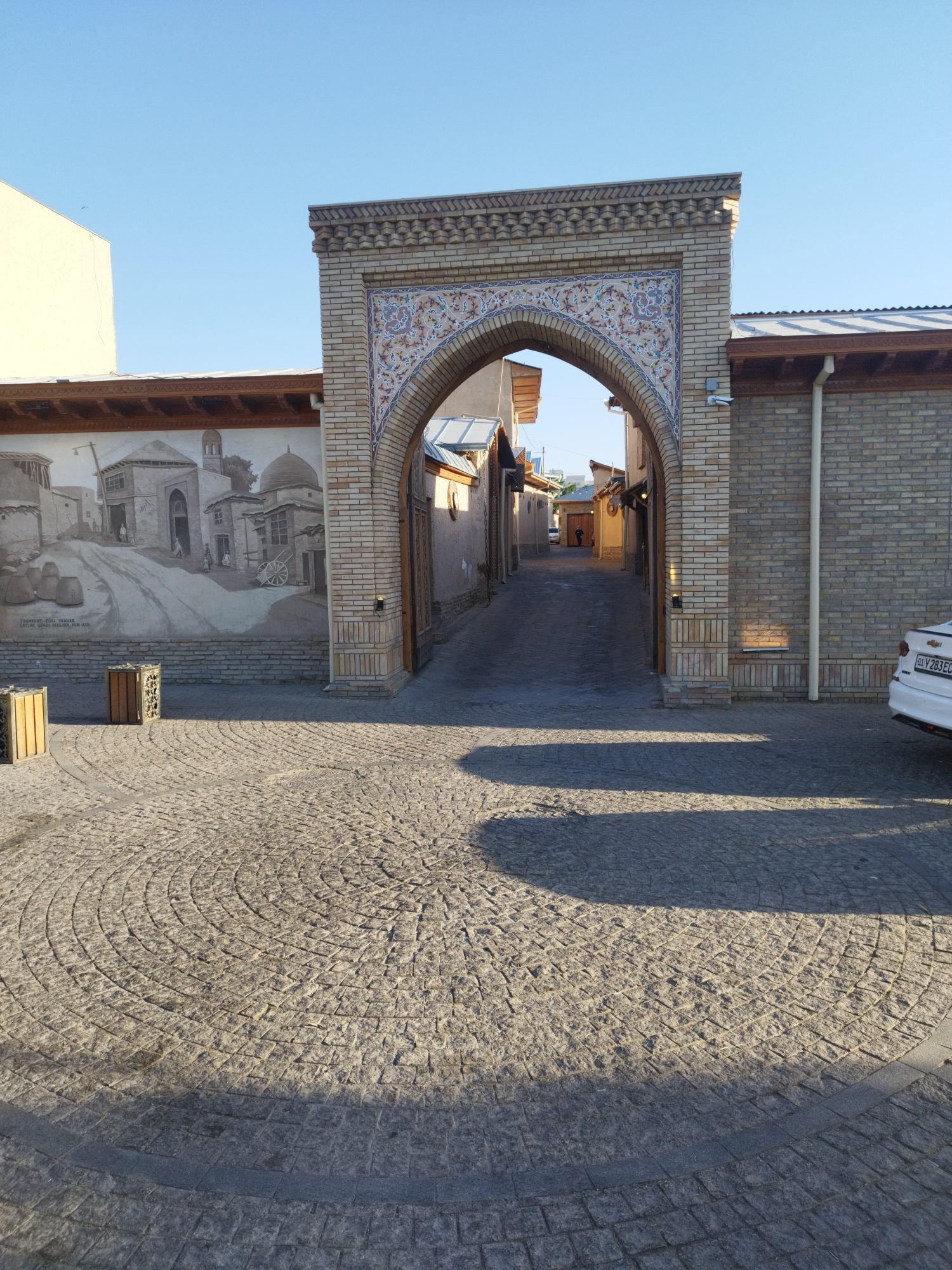 An outdoor, eye-level, medium shot captures a sunlit, arched brick entryway into a narrow street. The entryway is adorned with intricate, colorful tilework in shades of blue, red, and yellow, set against a pale brick facade. To the left of the entryway is a large mural depicting a historical street scene with buildings and figures, painted in monochromatic tones. The foreground features a circular cobblestone patio with a concentric pattern. Two wooden trash receptacles with decorative cutouts are positioned on the patio. A white car is partially visible on the right side of the frame, parked near the brick wall. The street beyond the archway is paved and lined with buildings, with a few figures walking in the distance. The sky above is clear and blue, casting shadows across the cobblestones.