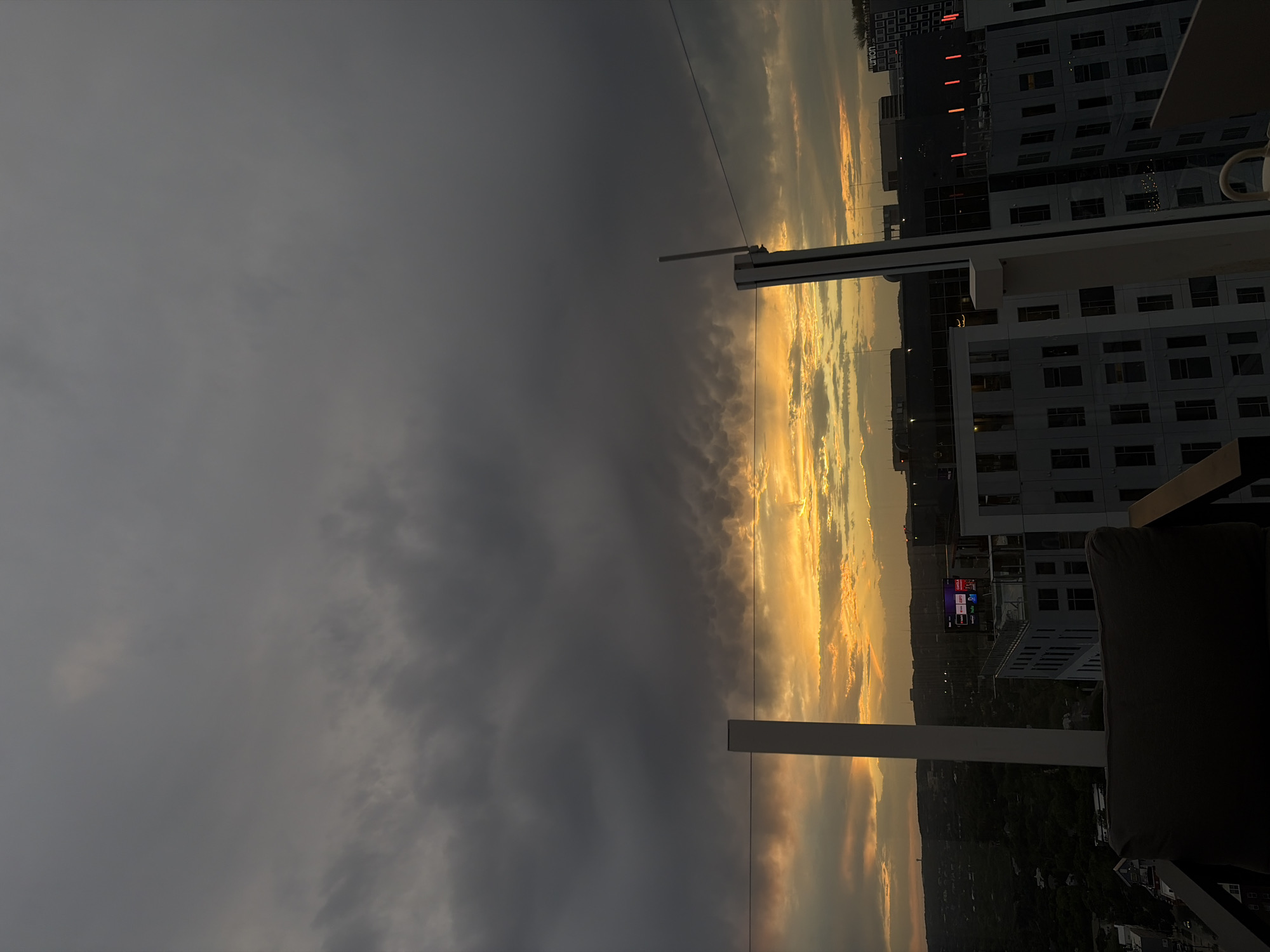 The image shows a sunset over the city of Austin, Texas, United States of America, as viewed from a high vantage point, likely a rooftop or high-rise balcony. The sky is divided between a dark, overcast area to the left, and a bright area with golden clouds to the right, where the sun is setting. Modern buildings with multiple windows are visible to the right, with the name "UNION" displayed on the side of one. A white structure is partially visible in the foreground. A portion of a chair is present in the lower right corner.