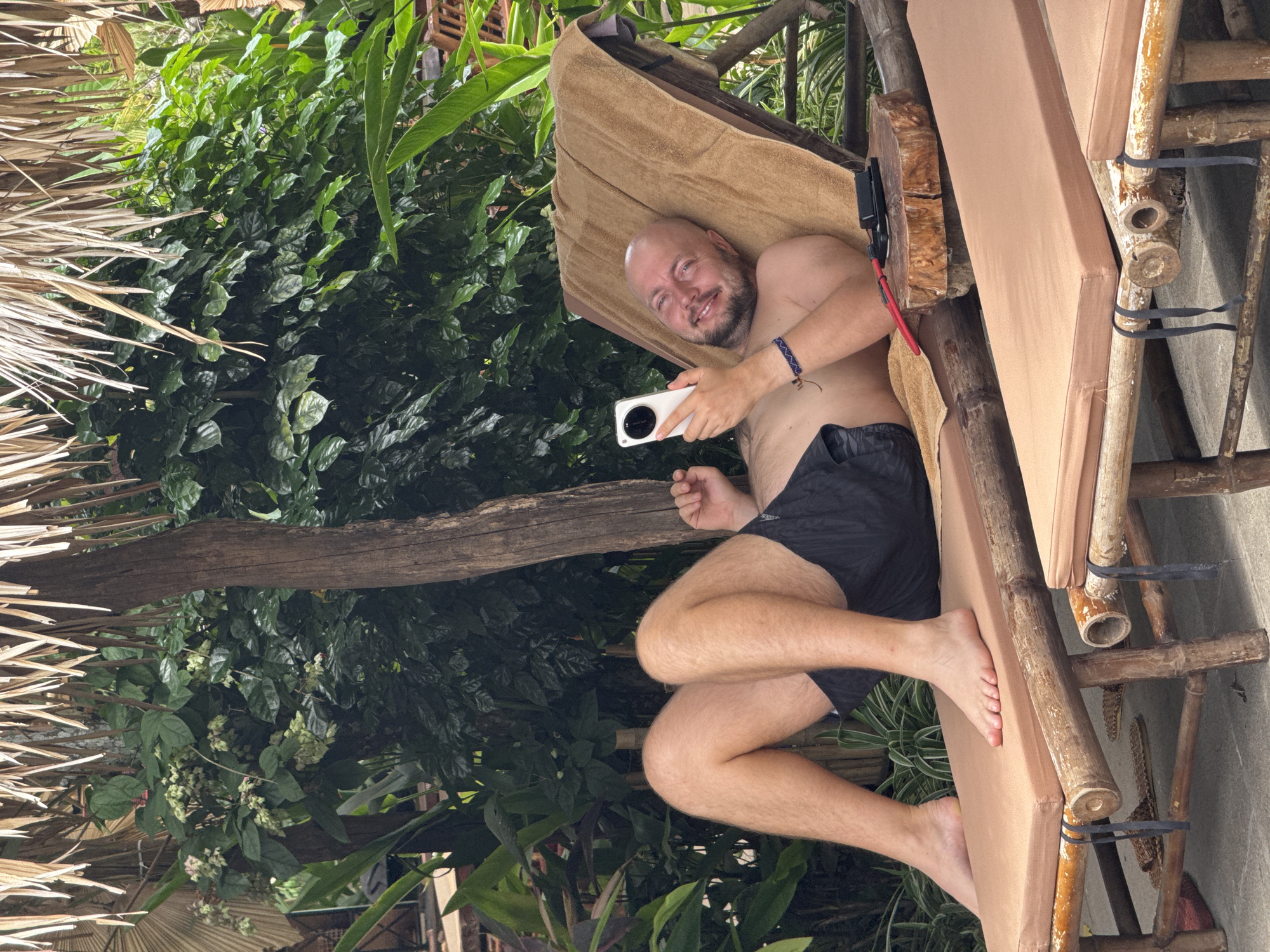 A bald, Caucasian man with a beard is reclining on a brown towel draped over a bamboo lounge chair. He is wearing black swim trunks and is holding a white smartphone in his right hand, looking at it. He is smiling. The lounge chair is made of bamboo and has a brown cushion. Next to the chair is a small wooden table with a black object and a red cord on it. The man is outdoors, with lush green foliage behind him and a thatched roof overhead. The scene appears to be in a tropical or subtropical environment. The location is identified as Nam Hoa Lư Ward, Vietnam.