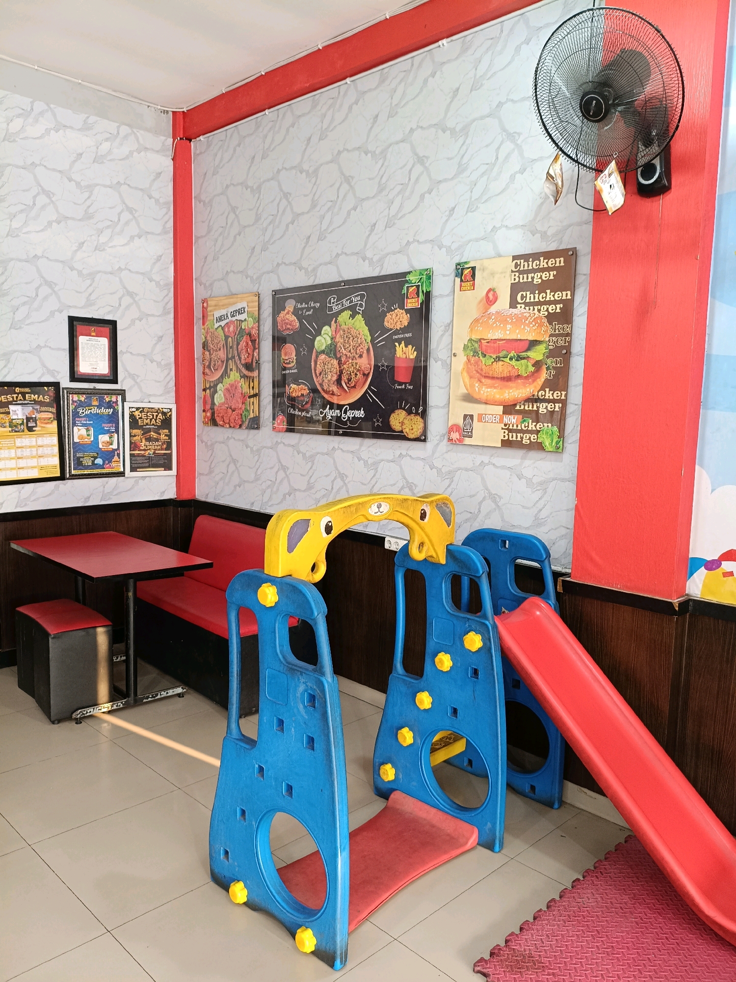 The image shows an indoor dining area, likely a restaurant, with a playful children's play structure in the foreground. The walls are covered in a light gray, marbled wallpaper and trimmed with red paint. On the walls are several framed posters advertising food, including chicken dishes and burgers. A large black oscillating fan is mounted high on the right wall. To the left, a red booth-style seat is visible, along with a small red table and a black and red stool. The floor is tiled in a light color. The children's play structure is made of bright blue plastic with yellow accents and features a red slide. The overall impression is a family-friendly establishment, possibly in Ngemplak, Indonesia, given the context. No people are visible in the image.
