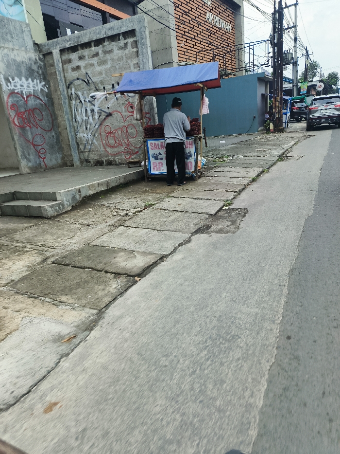 A man is standing in front of a street food stand, located on the sidewalk beside a paved road. The stand has a blue awning and is stocked with red fruit. The man is wearing a long-sleeved shirt and pants. He is facing the stand. The stand is located near a building with graffiti on its exterior. There is a blue door behind the stand. The road is empty, but there are parked cars in the background. The sidewalk has a concrete surface with patches of grass. The location is in Ngamprah, Indonesia, based on the coordinates (latitude: -6.868, longitude: 107.519).
