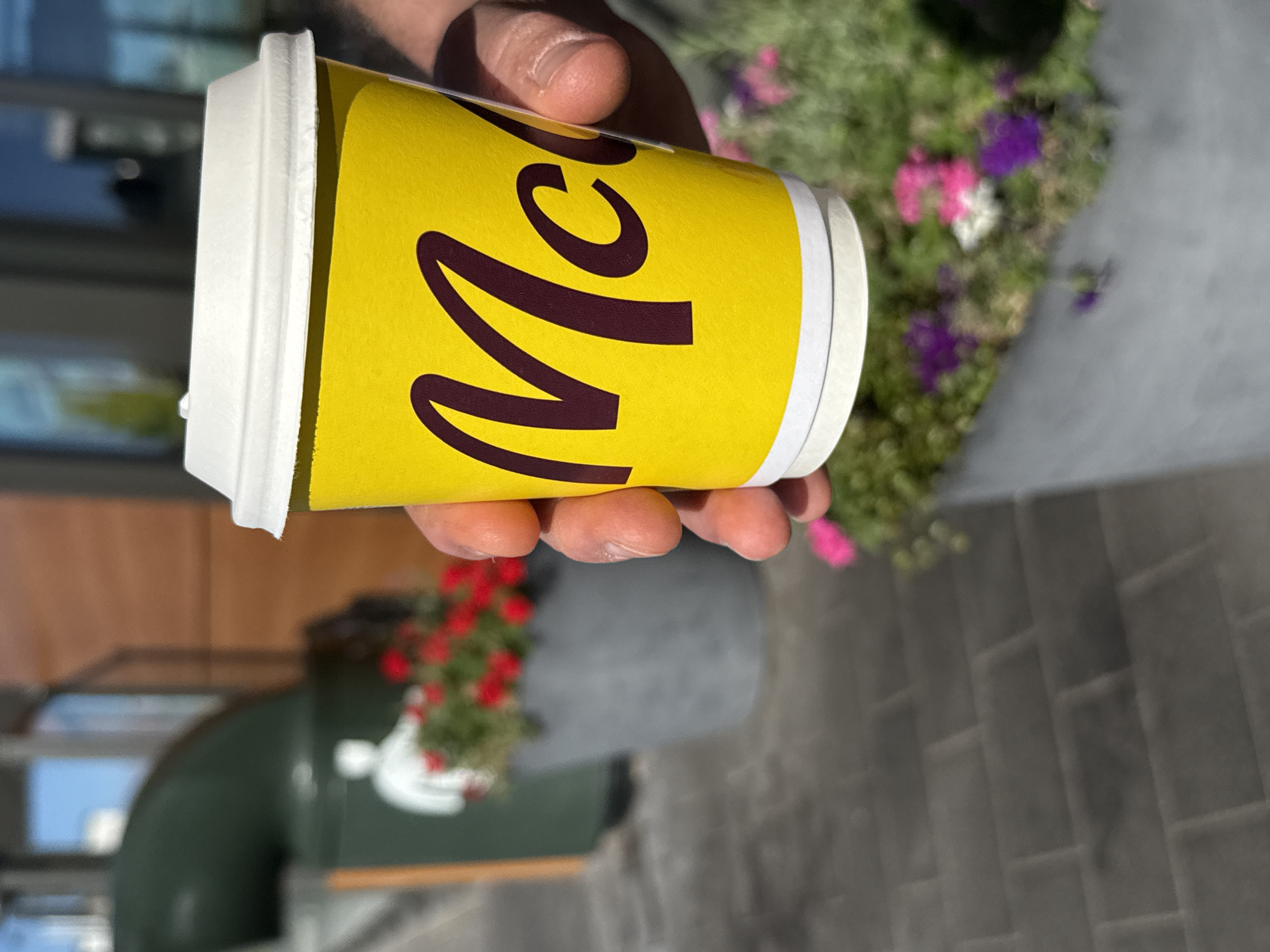 A close-up shot shows a person's hand holding a yellow McDonald's coffee cup with the "Mc" logo visible. The cup has a white lid and a white band around the bottom. In the background, out of focus, are a green trash receptacle with a white silhouette of a person on it, a planter with red flowers, and another planter with purple and pink flowers. The ground is paved with grey rectangular bricks. The scene appears to be outdoors, possibly near a McDonald's restaurant. No specific details indicate this location is in Werther, Germany.