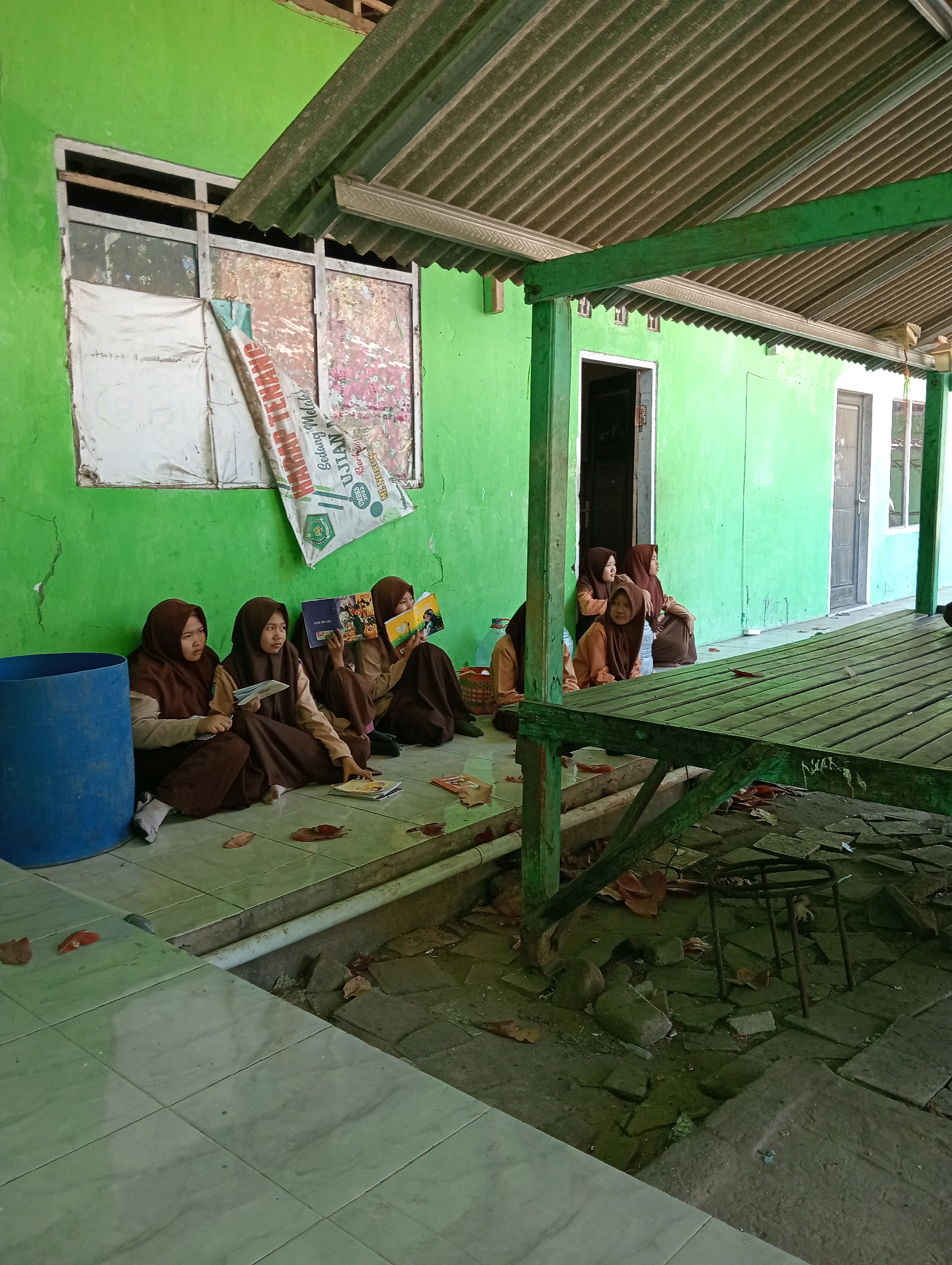In the city of Pagerungan Kecil, Indonesia, a group of four young female students, dressed in brown hijabs and uniforms, are sitting on a tiled floor outside a building with bright green walls. Two students are reading books, one of whom is holding an open book and looking towards the viewer. Another student is holding a stack of papers. The fourth student is looking away from the camera. To their left is a large blue barrel. In front of them, on the floor, are several fallen leaves and some books. Above the students, a corrugated metal roof is supported by green-painted wooden beams, casting a shadow over the scene. In the background, there is a window covered with torn plastic sheeting and wallpaper. To the right, a wooden structure with a slatted surface is visible, also supported by green beams. The environment appears to be an outdoor area adjacent to a building, possibly a school or community center.