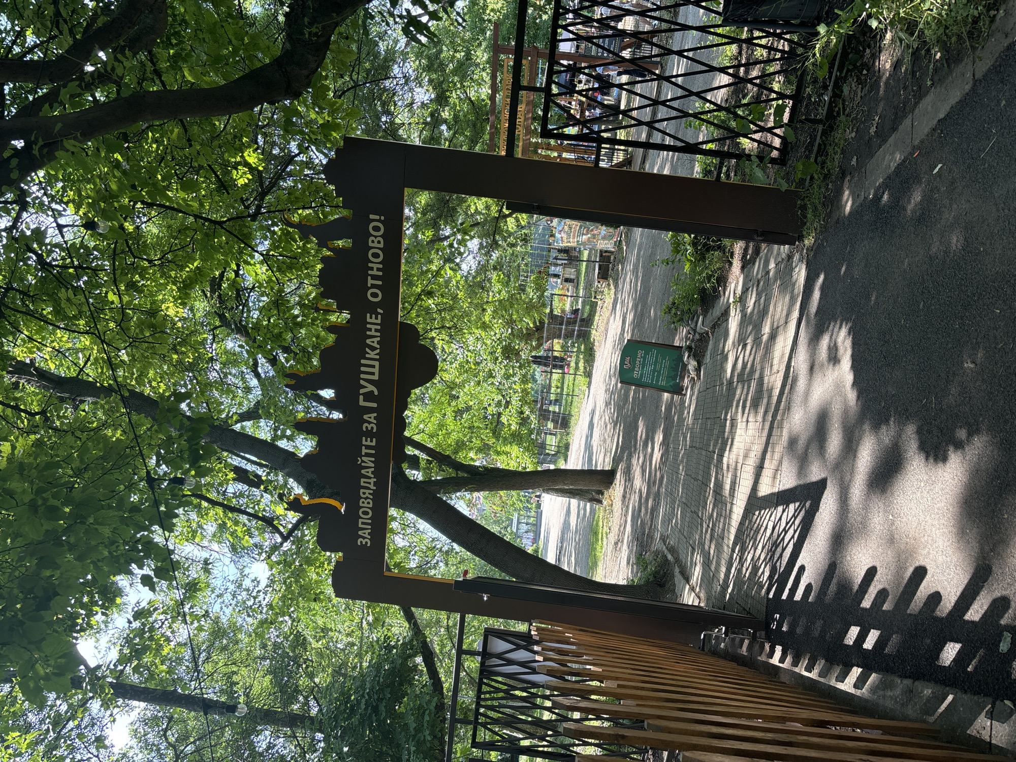 The image depicts an entrance to a park on a sunny day in Sofia, Bulgaria, specifically within Borisova Gradina. A large, dark brown archway serves as the main entrance, featuring a stylized, decorative top with yellow accents. The archway bears white Cyrillic text: "ЗАПОВЯДАЙТЕ ЗА ГУШКАНЕ, ОТНОВО!", which translates to "Welcome for a hug, again!" or "Come for a hug, again!".
To the left of the archway, a light brown wooden picket fence runs parallel to a paved pathway of rectangular bricks. To the right, a black metal fence with a diamond lattice pattern separates the path from a grassy area. The ground transitions from brick paving near the archway to dark asphalt further into the park. Sunlight casts prominent, elongated shadows from both fences and trees onto the ground, indicating a low sun angle.
Lush green foliage from overhead trees dominates the upper portion of the image, with sunlight filtering through the leaves and patches of bright blue sky visible. Several bare light bulbs hang suspended from wires between the trees.
A small, rectangular green sign is visible on the ground to the right of the left archway support, displaying partial Cyrillic text, "ГУШ... ОТВОРЕНО", likely indicating "Gushkane - Open", referencing the "Tree Hugging" area. In the background, a tree-lined path leads deeper into the park, where a few indistinct human figures can be observed walking in the distance. The overall scene suggests a public park environment designed for recreation.