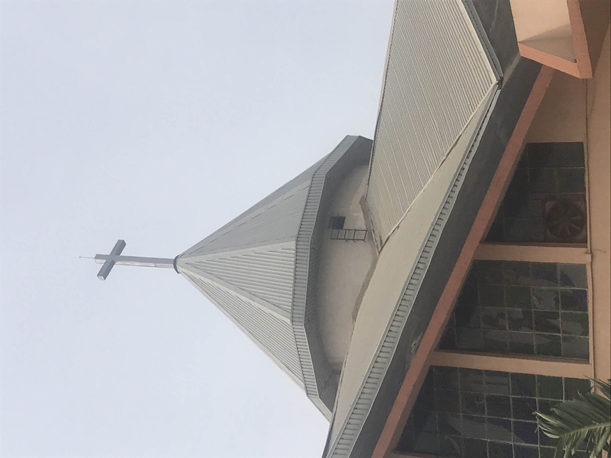 The image captures the upper section of a modern church building in Onitsha, Nigeria, viewed from a low angle against a bright, hazy sky. The central subject is a tall, multi-faceted spire, clad in ribbed, possibly corrugated metallic panels, that tapers sharply upwards to a minimalist cross at its apex. A small, fixed ladder is visible on one side of the spire, suggesting access for maintenance.
To the right of the spire, a portion of the church's main structure is visible. This section features architectural elements in a light, possibly peach-toned color, framing rectangular windows with dark, patterned panes that resemble stained glass or tinted blocks. The roofing of this adjacent section also appears to be corrugated metal, matching the spire. The overall architectural style suggests a contemporary design.
The scene depicts a static, architectural view under an overcast or diffused light sky, indicating it is likely daytime. No people are present, and there are no specific activities taking place. No visible text is discernible on the building.