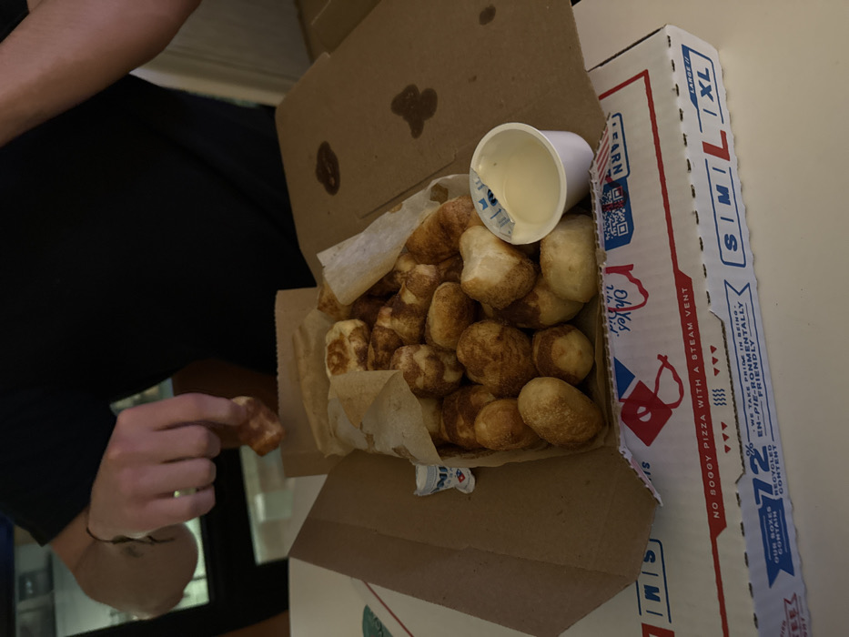 The image depicts an indoor scene, presumably in Boston, Massachusetts, United States, showcasing a Domino's Pizza box containing Cinnamon Bread Twists. A person, wearing a dark short-sleeved shirt and a dark bracelet on their left wrist, is visible, with their right hand actively retrieving one of the golden-brown, cinnamon-sugar-coated bread twists from the box.
The open, white cardboard Domino's box, displaying red and blue branding, rests on a light-colored, flat surface. Visible text on the box includes "NO SOGGY PIZZA WITH A STEAM VENT" and "OUR BOXES CONTAIN 72% RECYCLED CONTENT," along with size indicators (S, M, L, XL). The inside flap of the box contains several irregular dark stains. Inside the box, a substantial quantity of Cinnamon Bread Twists is piled within a brown paper liner. A small white plastic cup of white icing, with its foil lid partially peeled, is positioned among the bread twists. A smaller, mostly obscured white cup with blue branding is visible to the left. The background is dimly lit and indistinct.