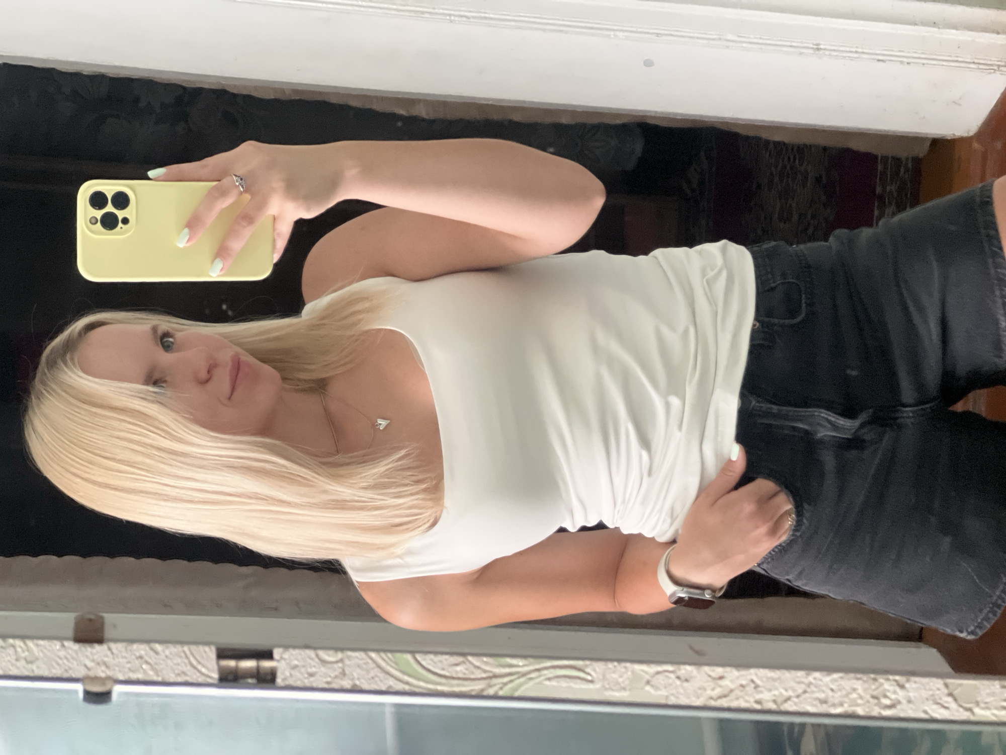 A young woman with blonde hair is taking a mirror selfie in a room. She is wearing a white tank top and black denim shorts. She has a light yellow phone case and a silver ring on her left hand, which is in her pocket. A silver watch is on her left wrist. She is wearing a heart-shaped necklace. The background shows a doorway and what appears to be a rug on the floor. The lighting suggests it is daytime.