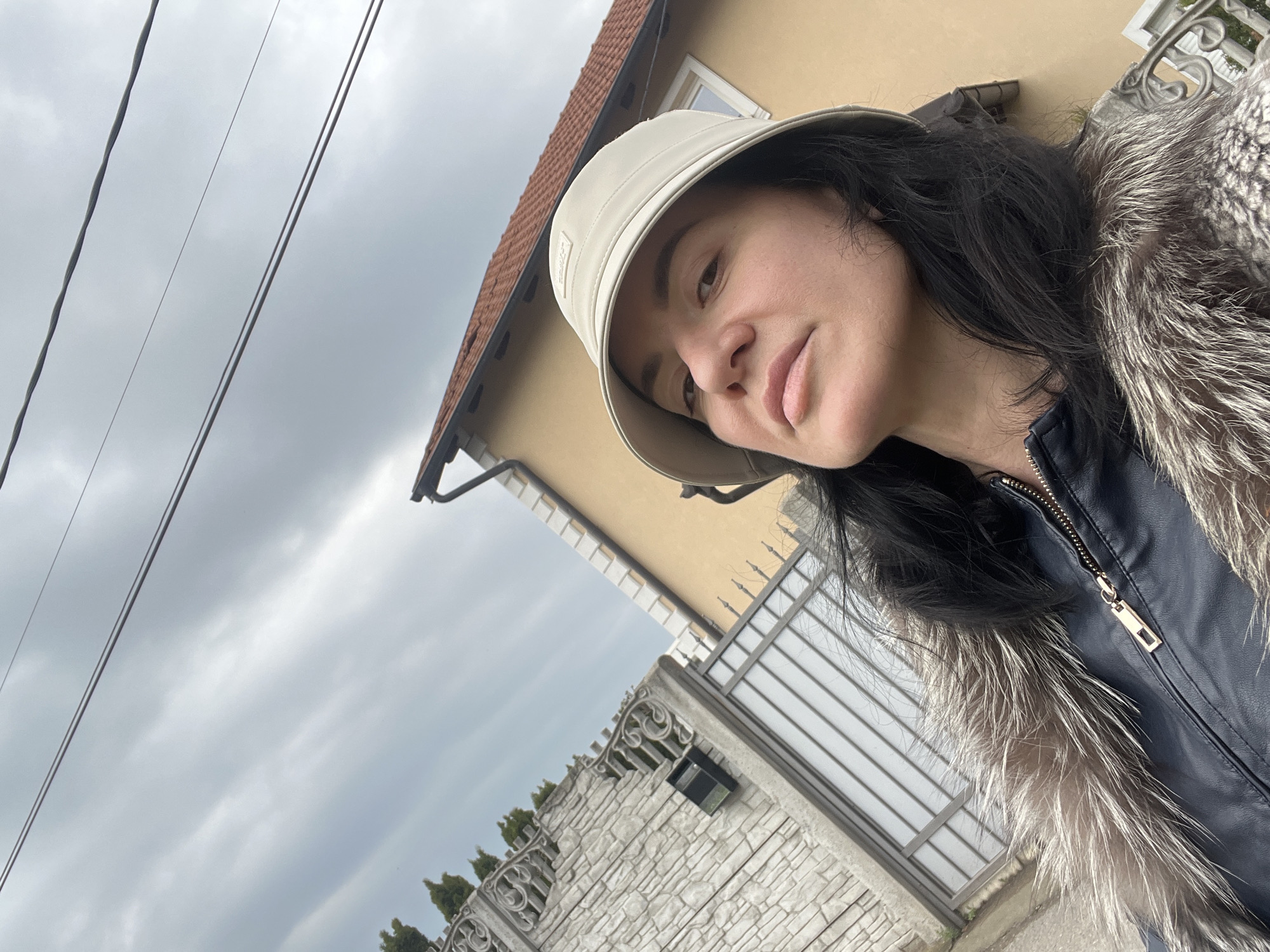 A woman with dark hair and a light-colored bucket hat poses for a selfie outdoors on an overcast day. She is wearing a dark jacket with fur trim around the collar. Behind her is a building with a yellow exterior, a reddish-brown tiled roof, and a white-framed window. A stone wall with decorative fencing and a metal gate are also visible. Power lines stretch across the sky in the upper left of the frame. The picture was taken in Belgrade, Serbia.