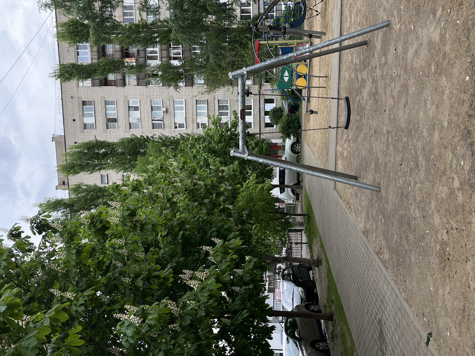 The image depicts a playground in an urban environment, potentially located in a residential area in the city of Unknown, Unknown. The foreground features a swing set made of metal poles with a single black seat hanging by chains. The ground surrounding the swing set appears to be covered in a sandy or gravel-like material. A paved walkway runs parallel to the playground area. In the background, there is a multi-story building with numerous windows, partially obscured by trees. Several cars are parked alongside the walkway, adding to the urban feel of the scene. The overall impression is one of a typical neighborhood park setting.