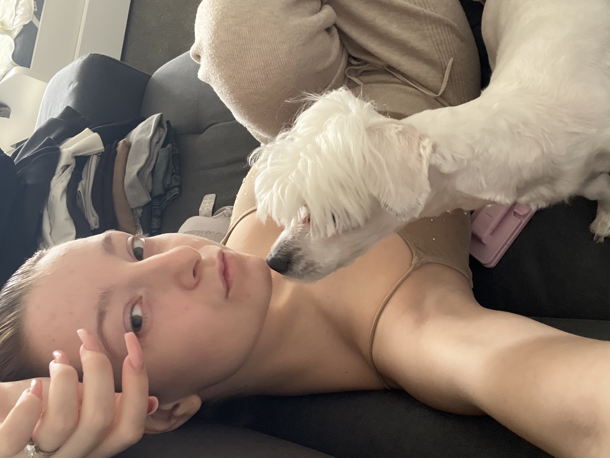 An indoor scene captures a close-up moment between a young woman and a small white dog. The woman lies on her back on a dark-colored couch, looking directly at the camera with a neutral expression. She has light skin, dark hair that appears slicked back, and is wearing a light beige or tan top with thin straps. Her left hand, adorned with long light-pink manicured nails and a ring on her ring finger, is near her face.
Above her head, a small, fluffy white dog, likely a Maltese or similar breed, leans down with its snout close to the woman's face, appearing to sniff or interact with her affectionately. The dog's fur is well-groomed.
In the background, to the left of the woman, a stack of folded clothes or textiles in various neutral tones (black, white, grey, brown) rests on the couch. Further back, a light-colored pillow or blanket and a white piece of furniture or shelf are visible. A pink object, possibly a phone or phone case, is partially seen near the dog on the couch. The lighting is soft, suggesting an indoor daytime setting. No visible text is present in the image.