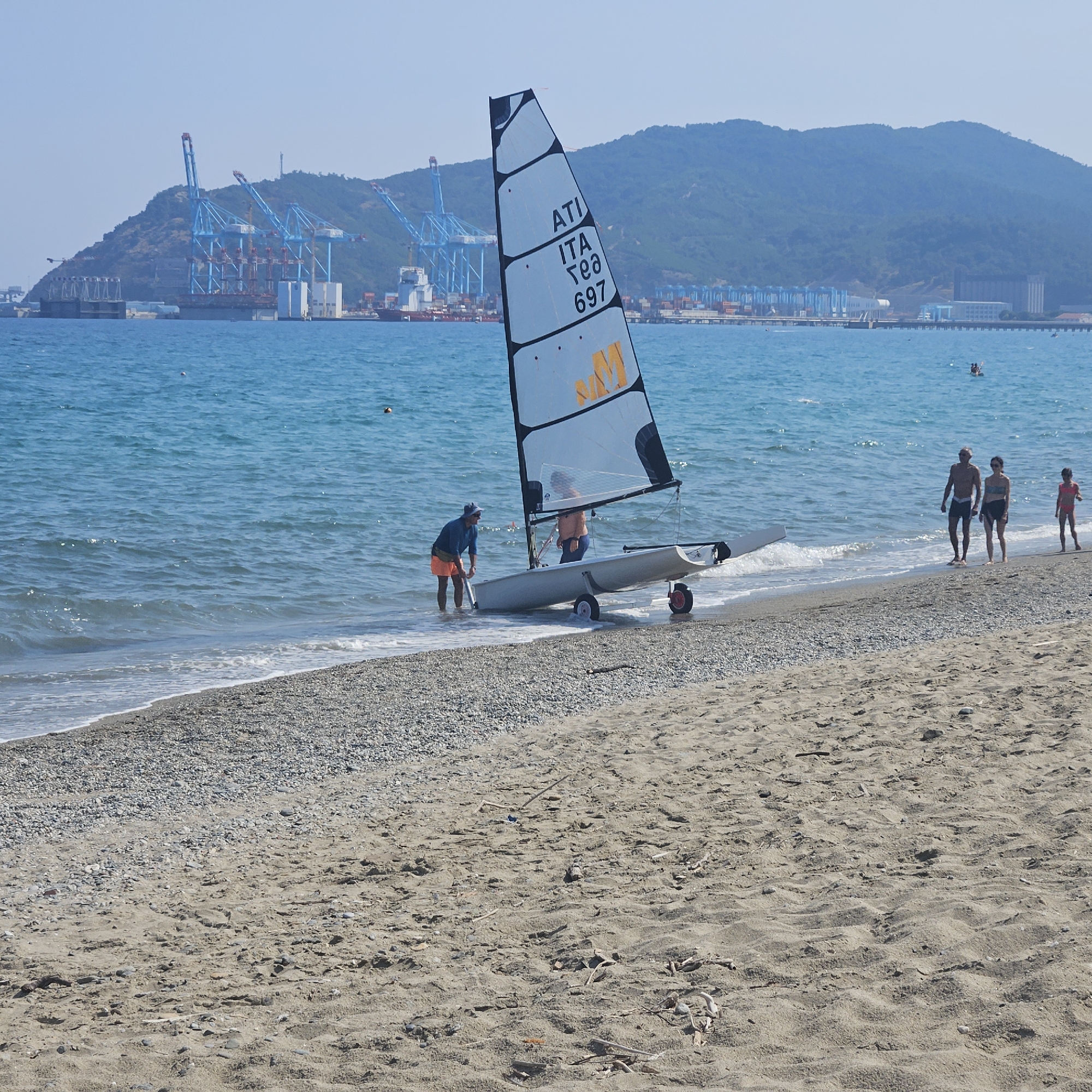 The image depicts a beach scene with a small sailboat being pushed towards the water by two individuals. The sailboat, a catamaran, has a white sail with black trim and the markings "ATI ITA Te8 697" and "MM" in yellow. One person, wearing a dark blue shirt and orange shorts, is in the shallow water next to the boat, appearing to push it. Another person, wearing a light orange shirt, is inside the boat. In the background, the turquoise ocean stretches to the horizon, with a mountainous landscape and a port complex featuring numerous cranes and shipping containers. A few other individuals are visible on the beach further down, including a couple and a child. The beach itself is a mix of sand and pebbles, with some driftwood scattered about. The sky is clear and blue. Based on the presence of the shipping port with its characteristic cranes and the mountainous coastline, the scene is consistent with the port area of Savona, Italy, specifically the industrial port facilities visible in the background.