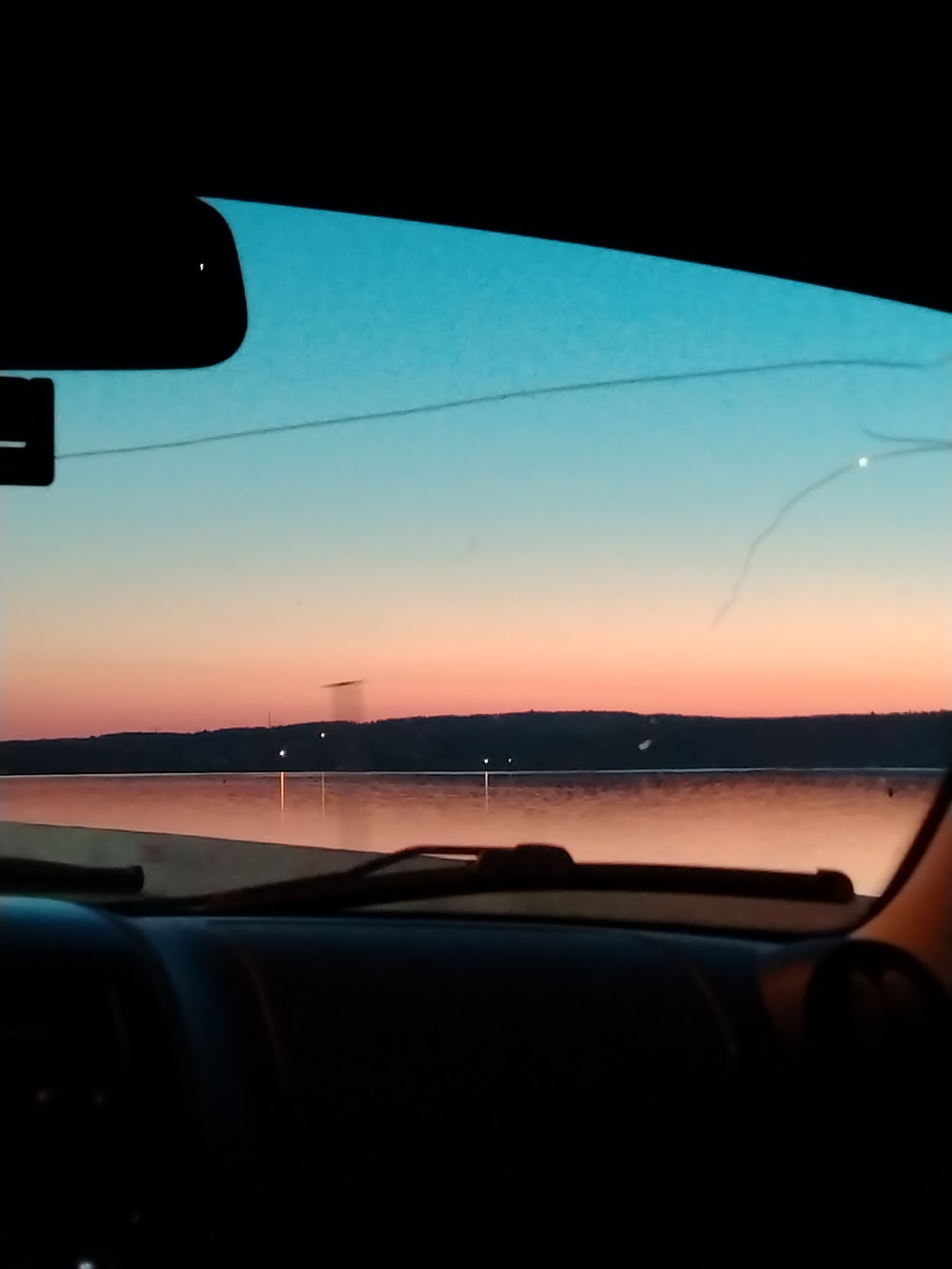 A photograph captures a view from inside a vehicle through a cracked windshield, looking out over a large body of water at either sunrise or sunset. The sky displays a gradient of colors, transitioning from a light turquoise blue at the top to soft pink and orange hues near the horizon. A dark, low-lying landmass, possibly a distant shore or hills, separates the colorful sky from the water. Several small, bright lights are visible on this distant landmass, with their reflections shimmering on the water's surface, which largely mirrors the pink and orange tones of the lower sky.
The upper left quadrant of the image shows a portion of the vehicle's dark rearview mirror. The windshield exhibits multiple fine cracks, prominently in the upper right section, and a distinct horizontal line stretches across its middle. The dark dashboard and the top of two windshield wipers are visible at the bottom of the frame, partially obscuring the immediate foreground of the water. No individuals are discernible in the scene. The image provides no explicit visual markers or landmarks to forensically confirm the location as the city of Chaikovsky, Russia.