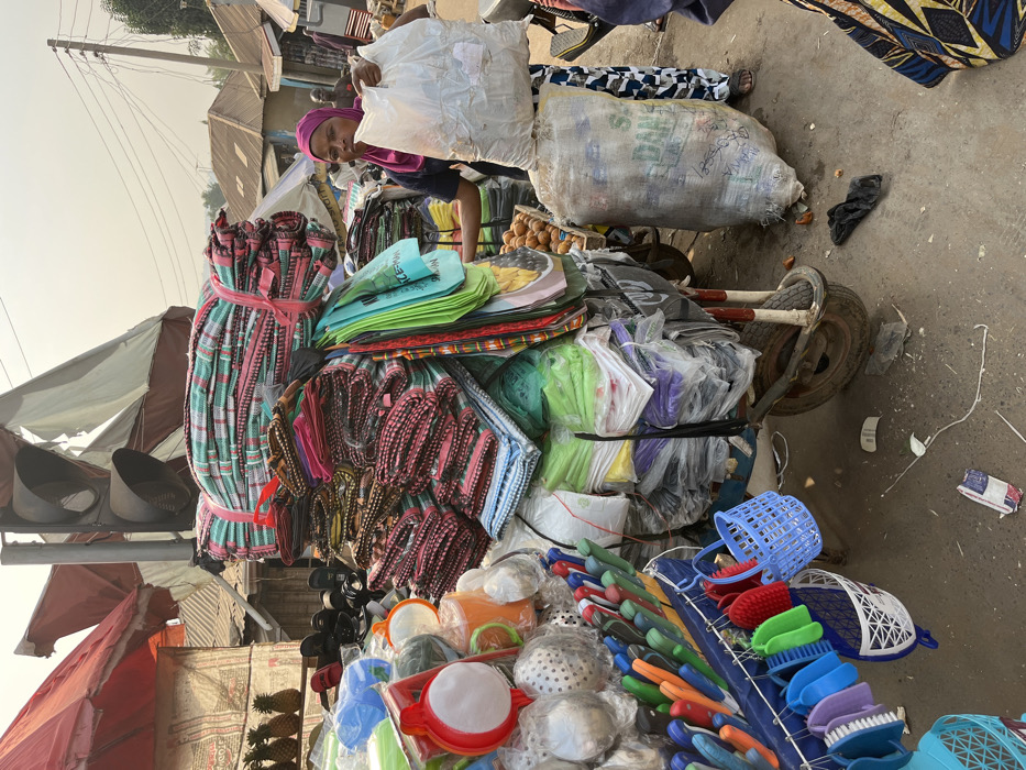 A person with a headscarf stands behind a cart piled high with goods. Bundles of tied textiles in red, white, and green, and stacks of colored shopping bags are visible. Below are household items like colanders, colorful brushes, and small plastic baskets. The cart appears to be loaded with a white sack and another large bag. The background features other stalls and structures. The photograph is taken outdoors in what appears to be a bustling marketplace.