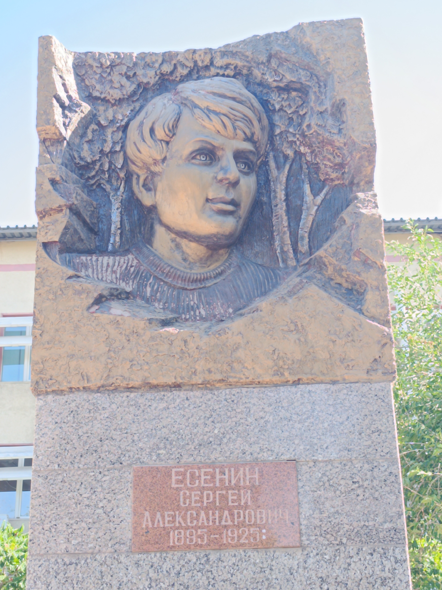 The image shows a monument featuring a bas-relief portrait of a young man, likely Sergei Yesenin, a renowned Russian poet. The portrait is carved into a textured, light brown stone slab, with trees or foliage depicted behind the subject's head and shoulders. The slab is mounted on a larger, grey, granite-like base. A red plaque is affixed to the base of the monument, inscribed with Cyrillic text that appears to read "ЕСЕНИН СЕРГЕЙ АЛЕКСАНДРОВИЧ 1895-1925". The monument is outdoors, with a clear blue sky in the background. To the left of the monument, a building with windows and a roof is visible. To the right, lush green foliage suggests the presence of trees or bushes. The monument is located in the city of Taraz, Kazakhstan, specifically in front of the city administration building.