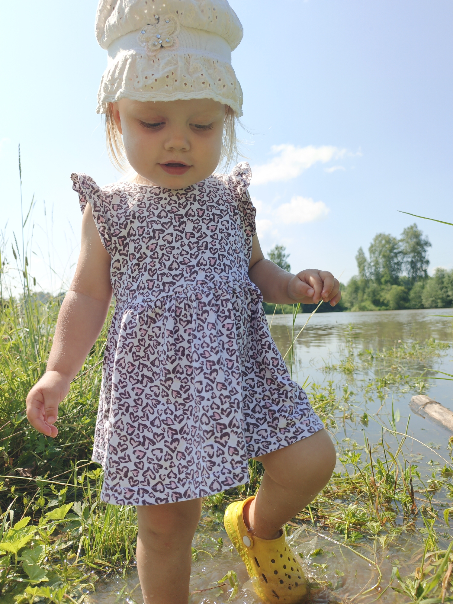 A young child wearing a white bonnet and a dress patterned with small pink and brown hearts stands in shallow water near the grassy bank of a river. The child's right leg is extended forward, with a yellow rubber shoe on their foot. The background shows a calm river surrounded by lush green vegetation and trees under a bright blue sky with scattered clouds. The scene is set in the village of Urtiushka, Russia.