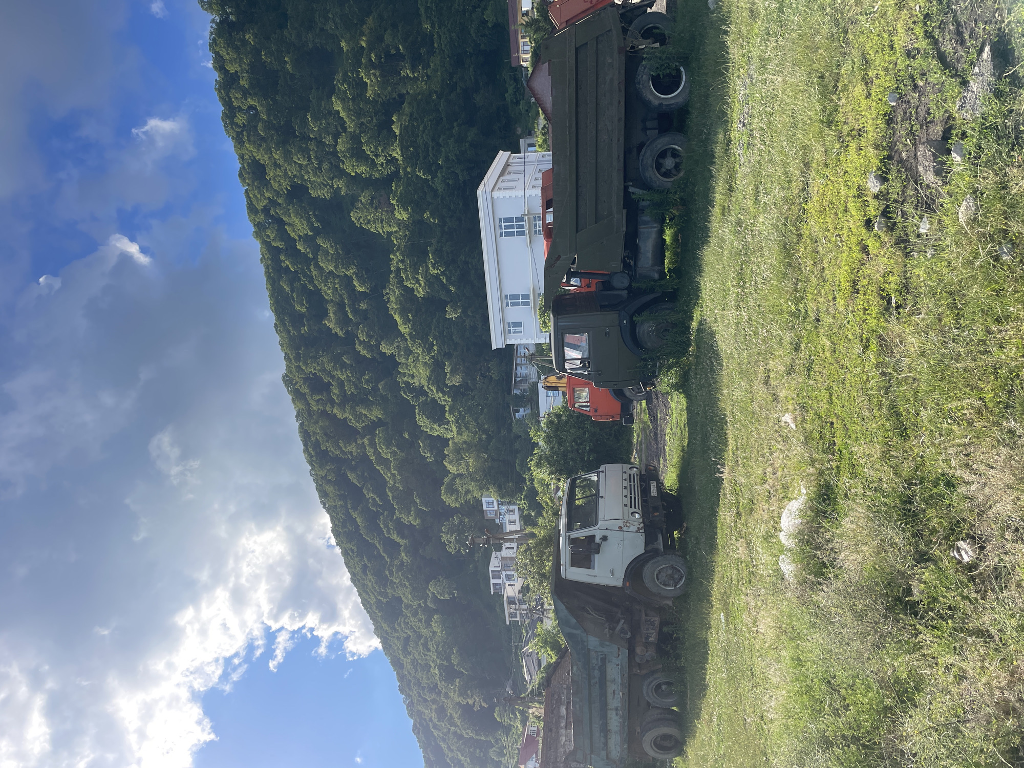 The image depicts an outdoor scene on a sunny day in Baranovka, Russia. In the foreground, two large dump trucks are parked on a grassy area. The truck on the left is lighter in color, while the one on the right is dark green. Behind the trucks, a densely forested green hillside dominates the background, with a few white buildings visible amidst the trees. The sky above is a mix of blue and clouds, with sunlight casting bright highlights. The environment appears to be a somewhat undeveloped area, possibly on the outskirts of a settlement, given the presence of the industrial vehicles and the natural landscape. There are no people visible in the image.